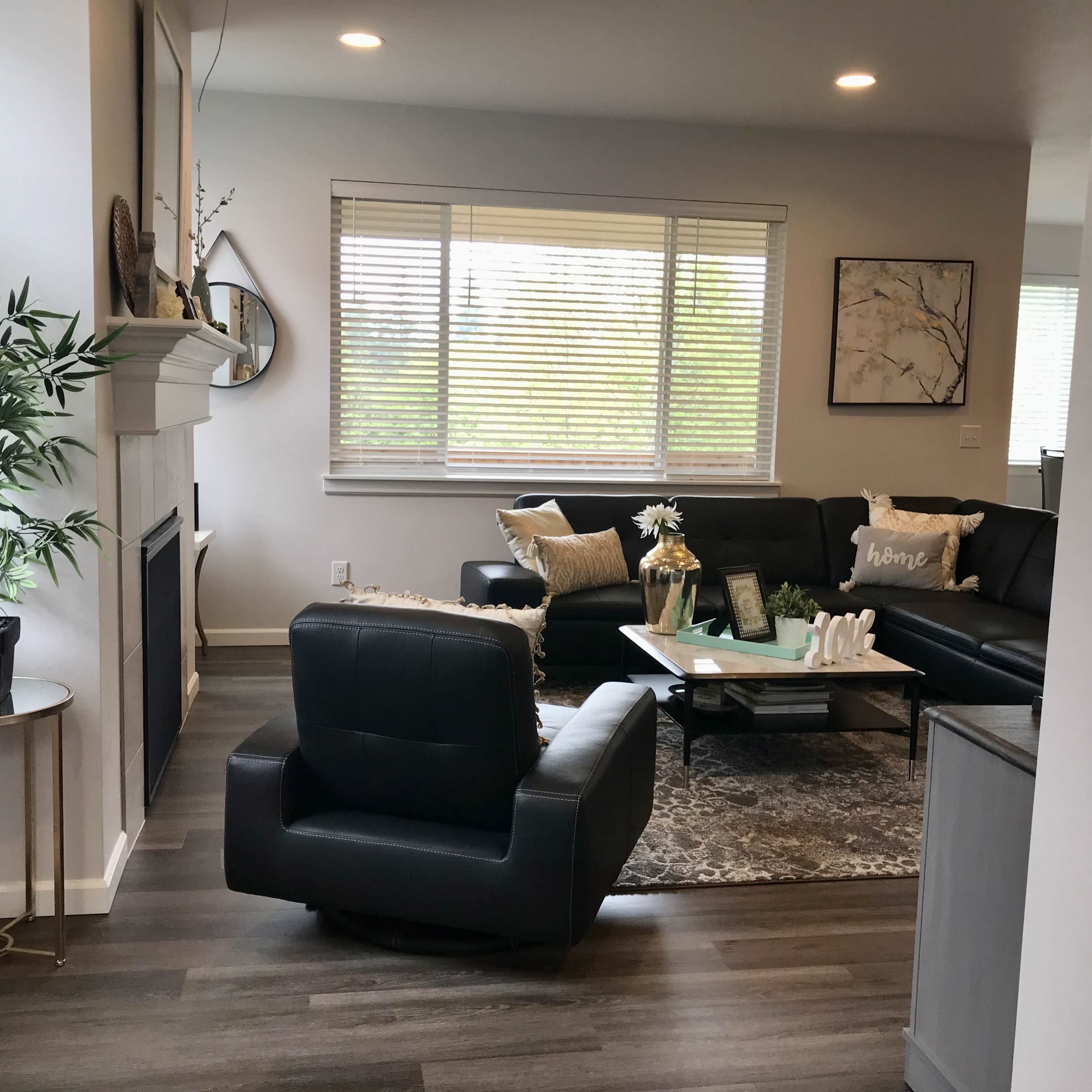 A modern living room features a black leather sofa, a swivel chair, a coffee table with decorative items, and large windows allowing natural light.