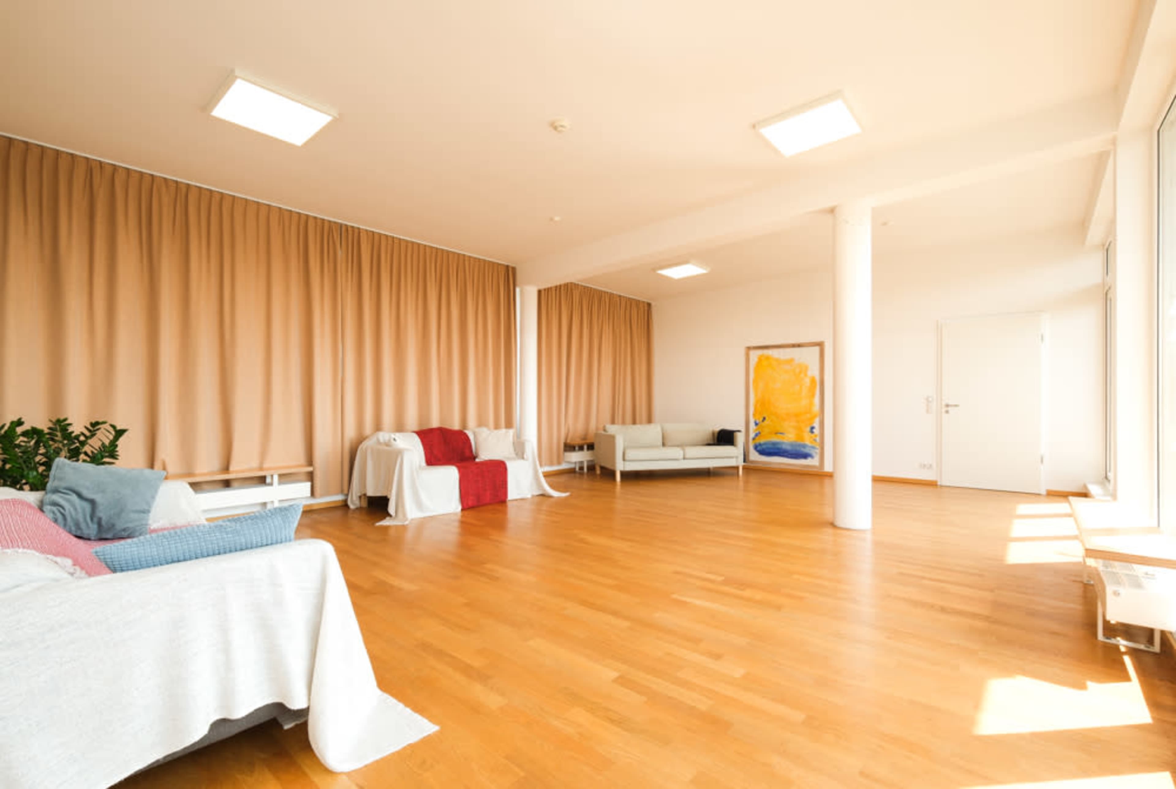 The image shows a spacious and well-lit room with wooden flooring, featuring two couches, light-colored curtains, and a colorful painting on the wall.