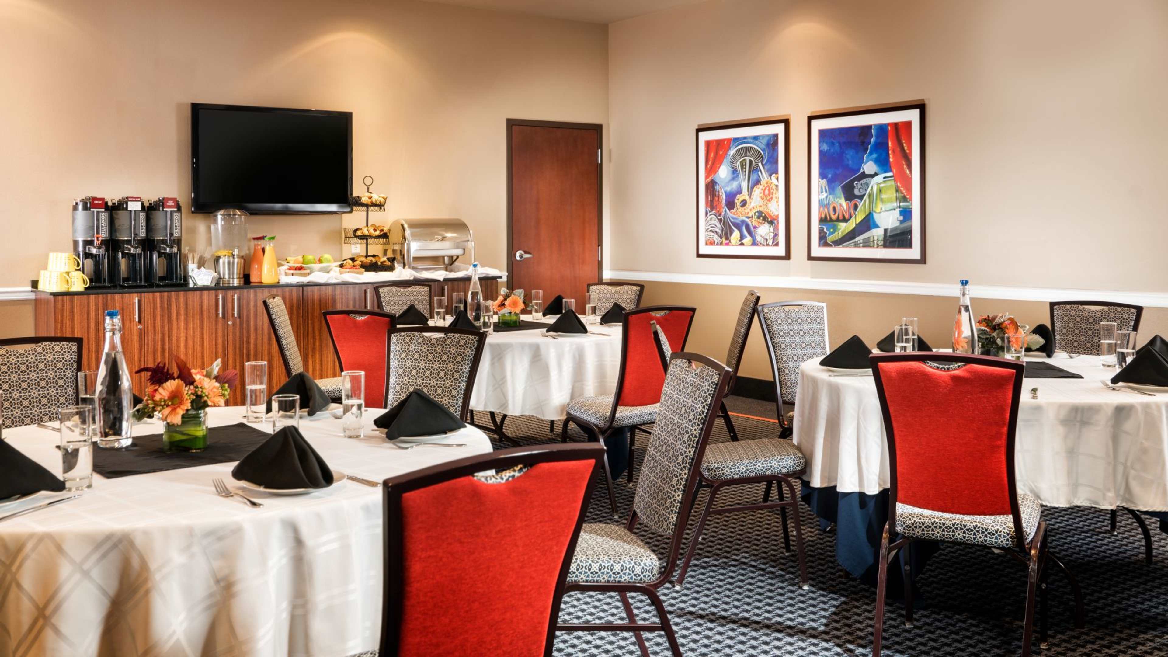 A neatly arranged dining room with round tables, red and patterned chairs, and a small buffet area in the background.