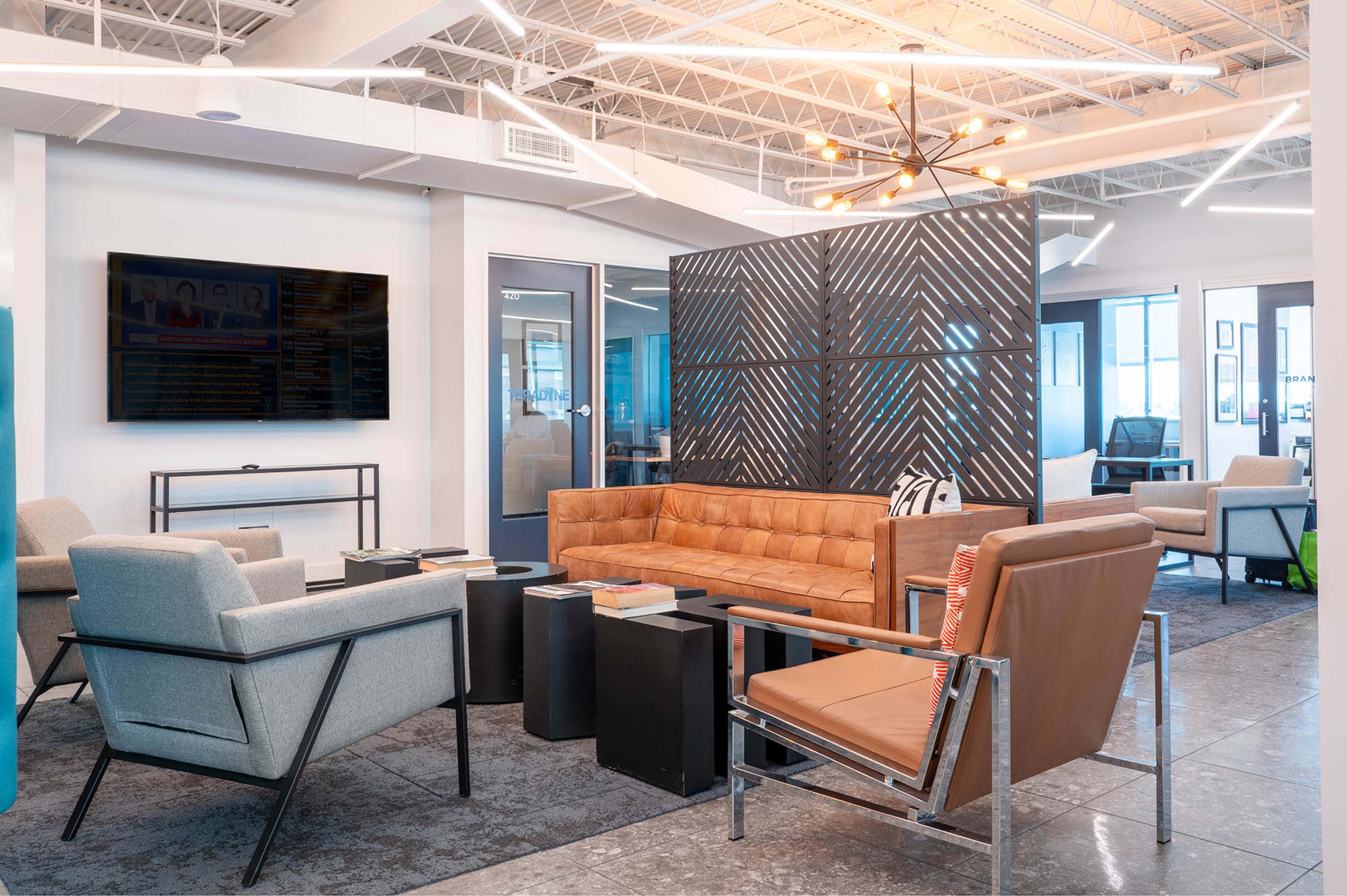 The image shows a modern office lounge with a mix of seating options, including a brown leather couch, gray armchairs, a coffee table, and a large screen on the wall.