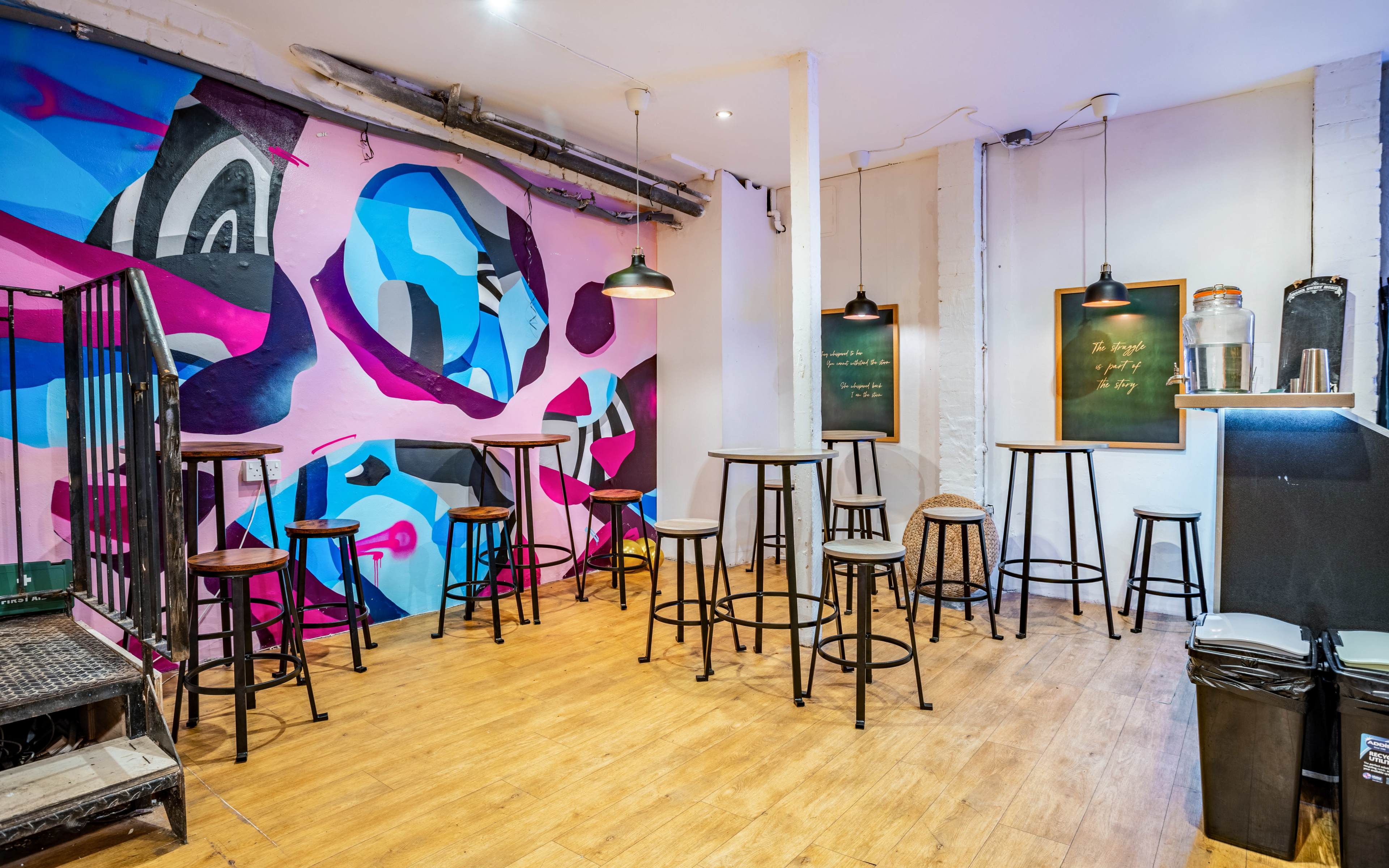 A room with wooden flooring, featuring several tall stools arranged around small tables and a colorful mural on the wall.