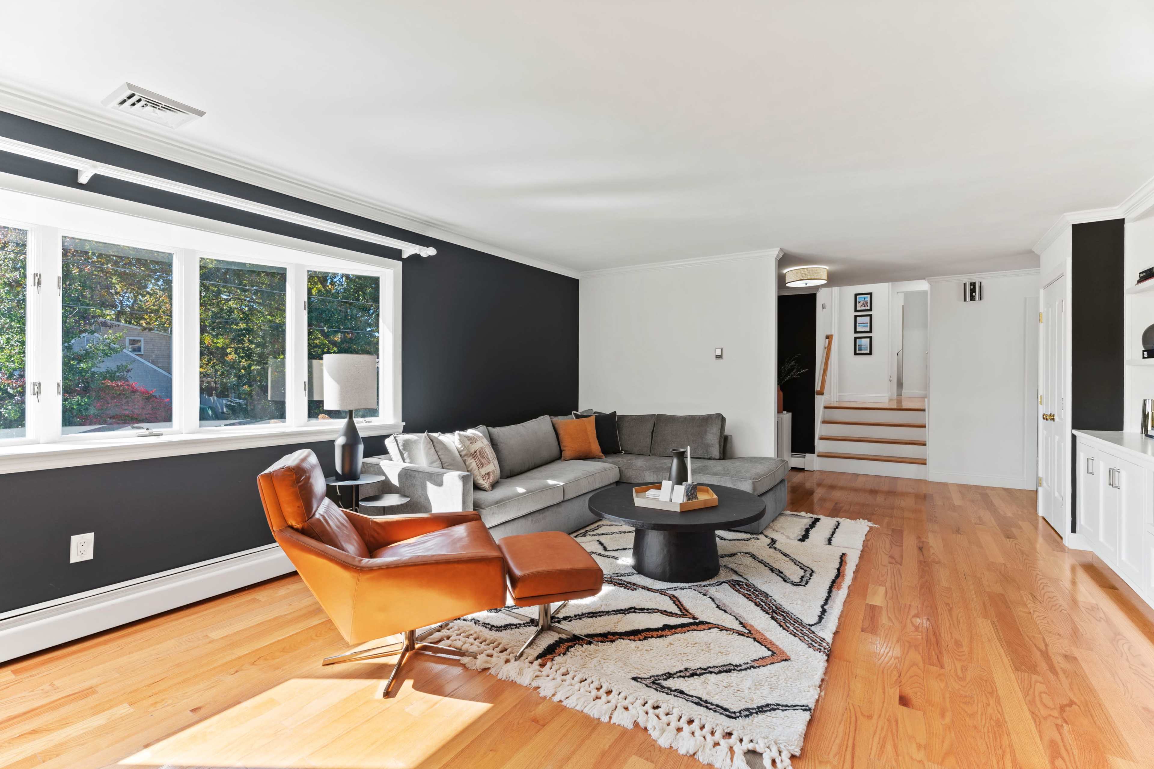 A living room features a gray sectional sofa, an orange accent chair, a circular coffee table, and large windows allowing natural light to enter.