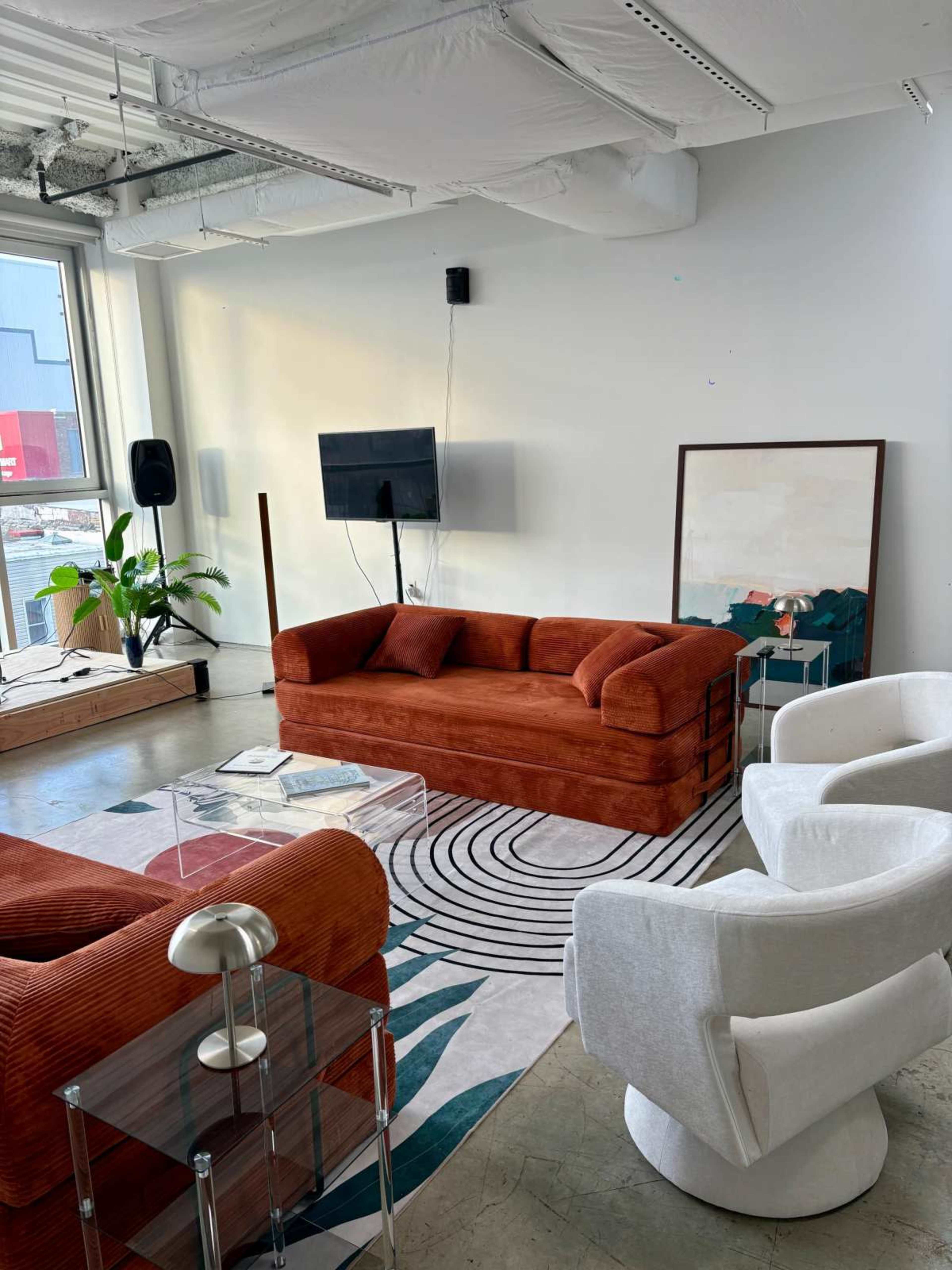 The image shows a modern living room featuring a red sofa, white armchairs, a patterned area rug, and a television mounted on the wall, with large windows allowing natural light to enter.