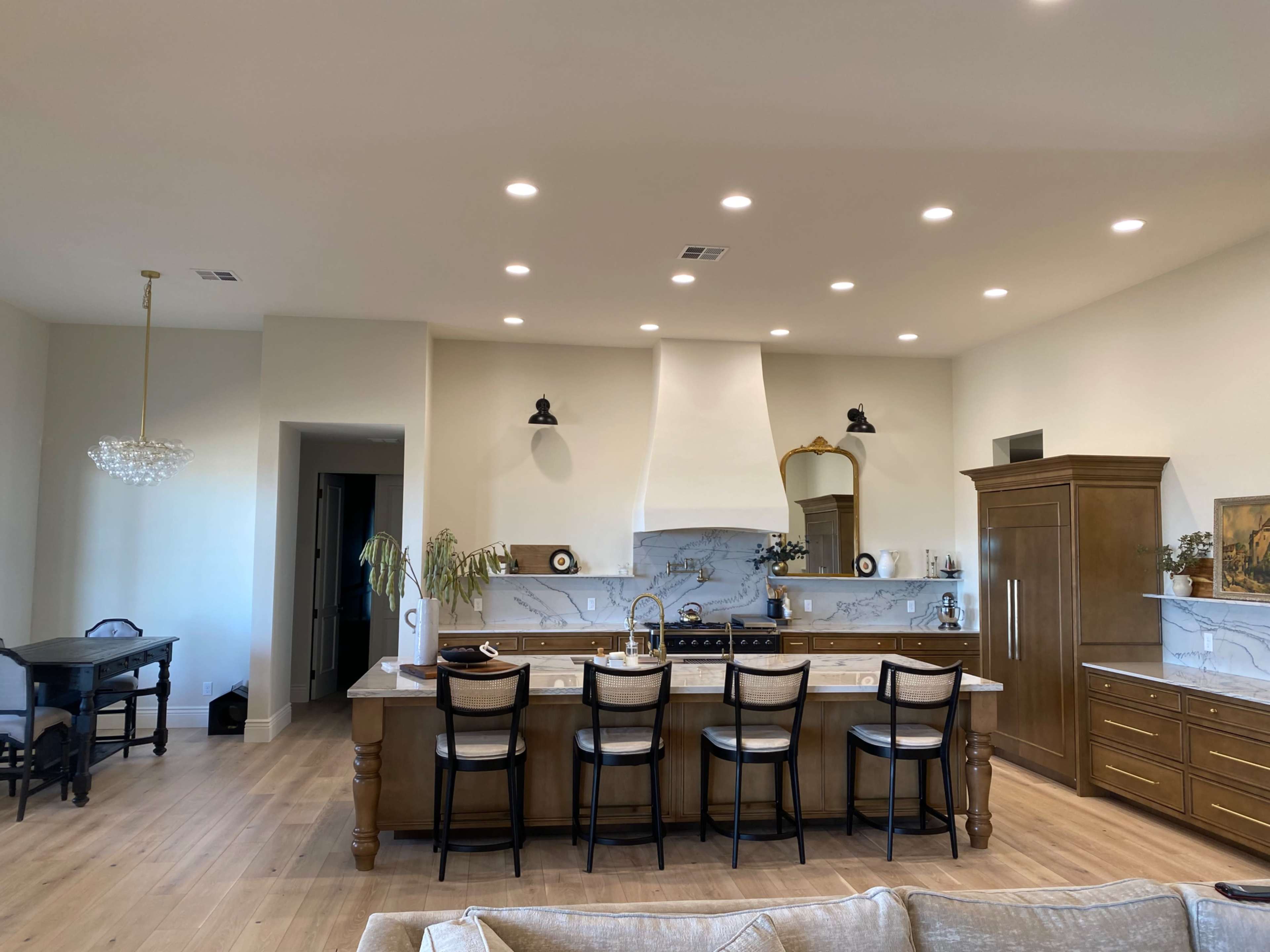 A modern kitchen features a large island with four bar stools, a white hood above the stove, and various decorative elements on the countertops.