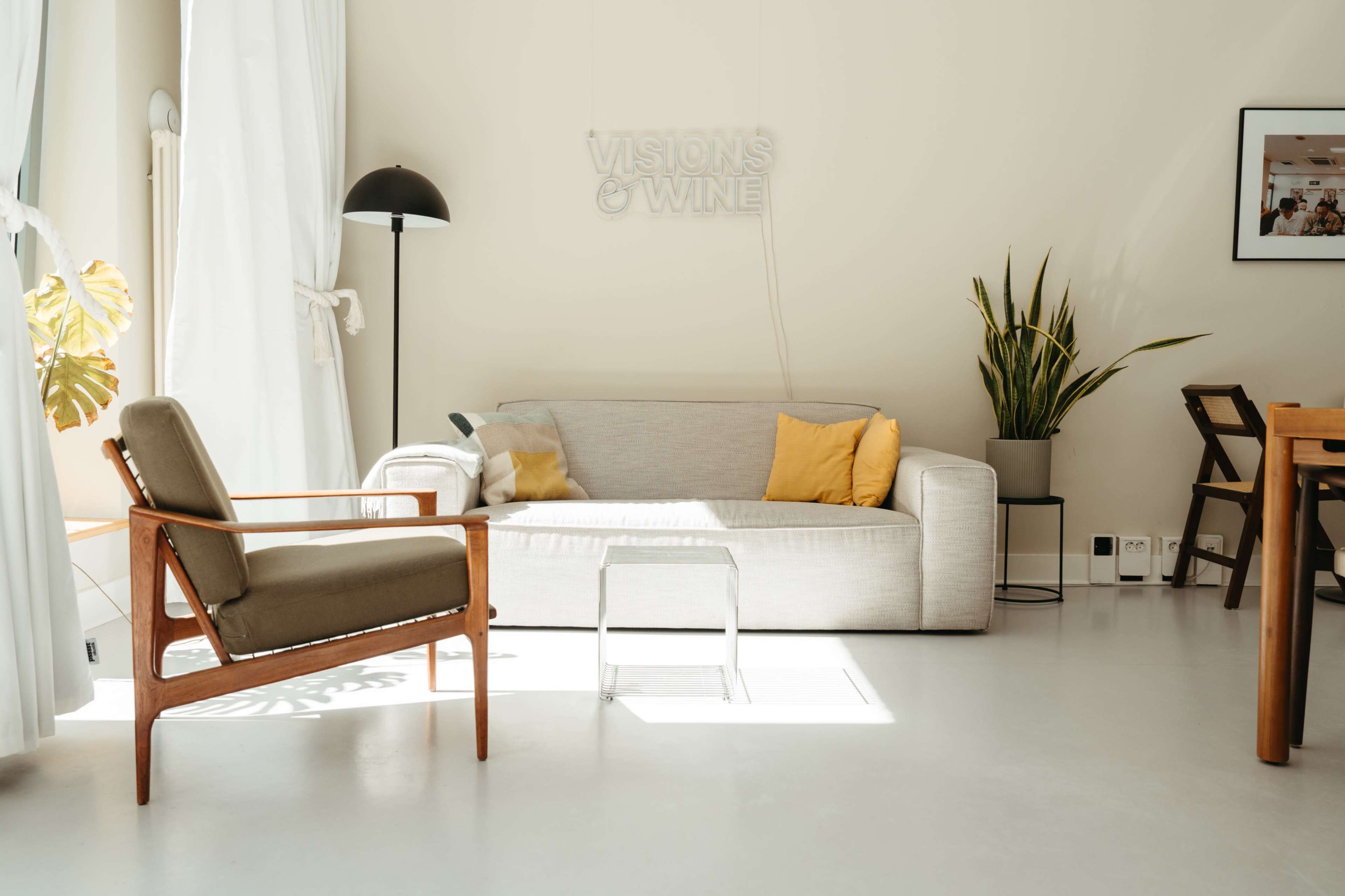 A minimalist living space features a light-colored sofa with yellow accent pillows, a wooden chair, a clear side table, and a potted plant.
