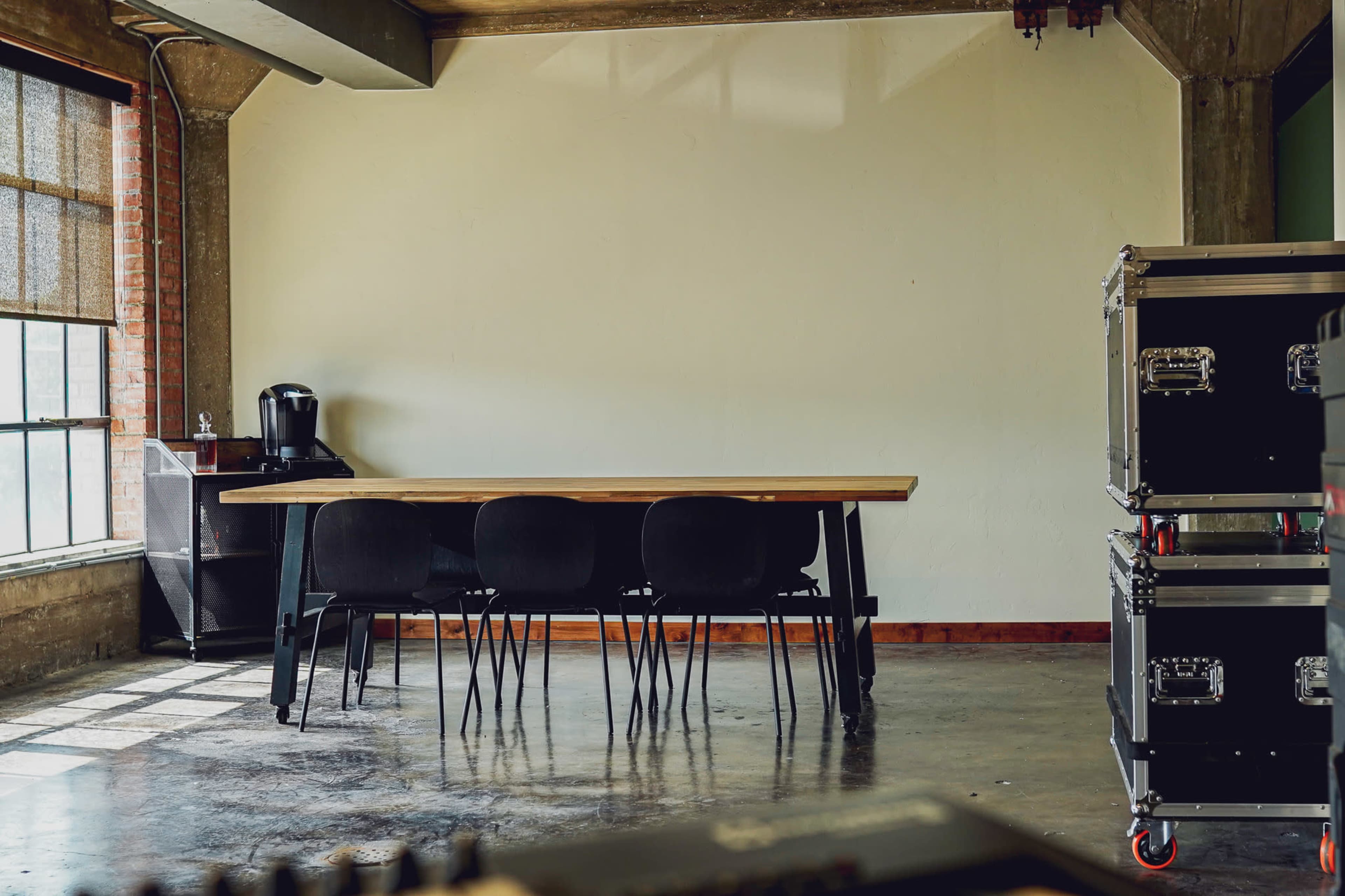 A long wooden table with six black chairs is placed against a plain wall in a room with large windows and industrial-style accents.