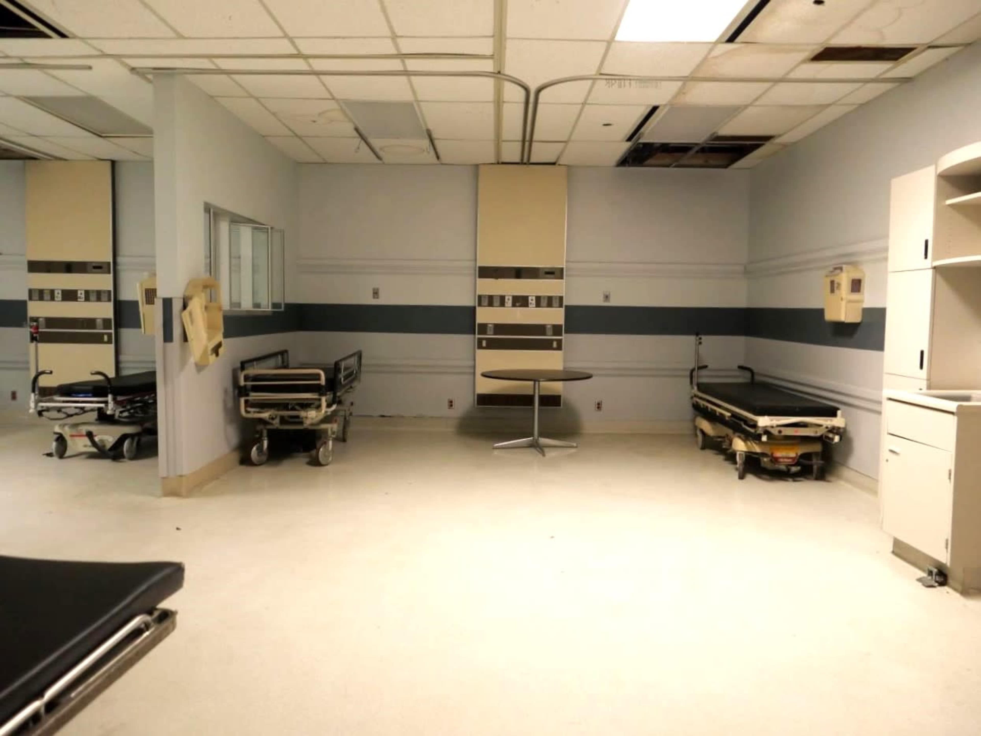 An empty hospital room features two hospital beds, a round table, and equipment storage against the walls.
