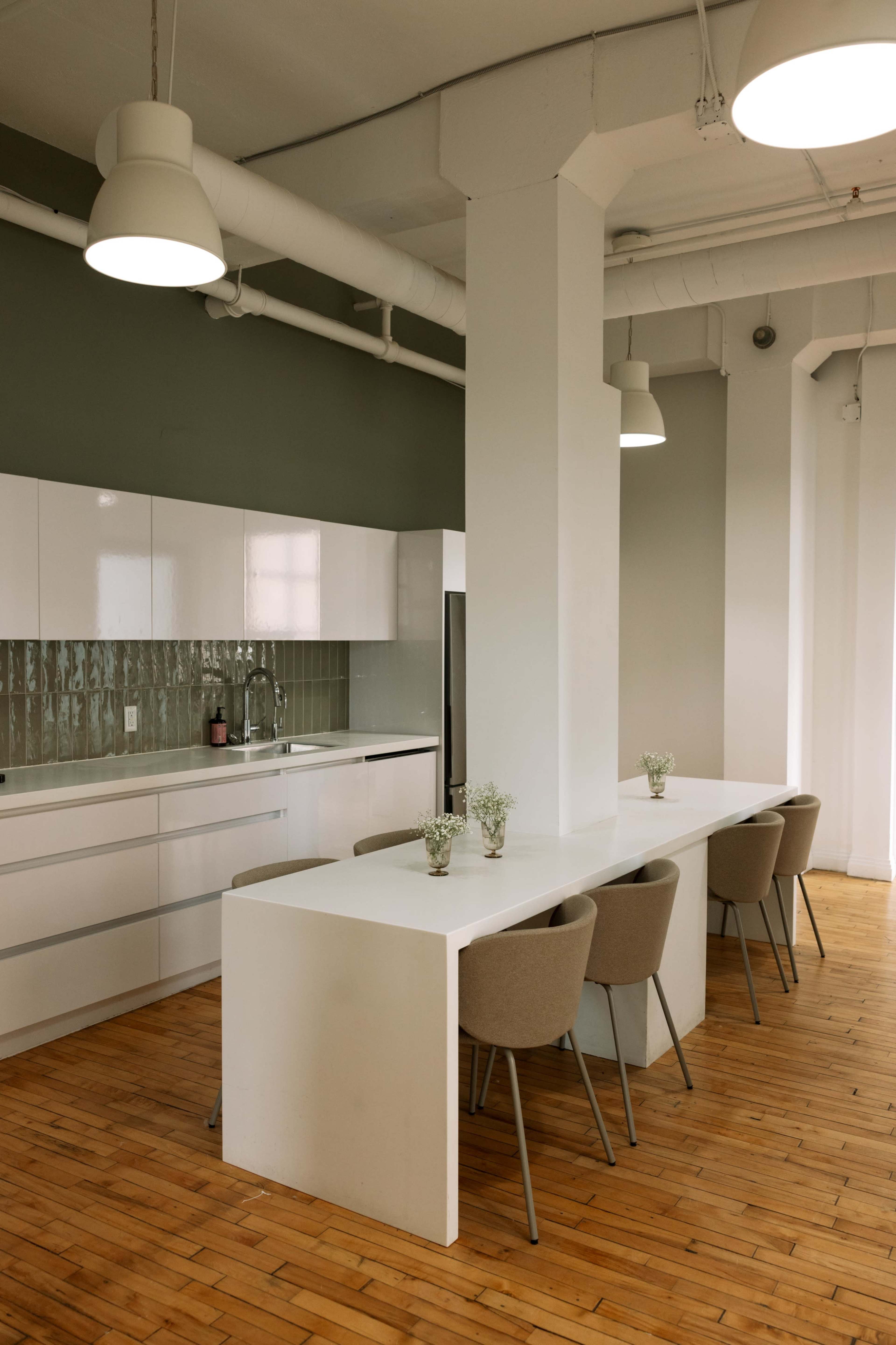 The image shows a modern kitchen area featuring white cabinetry, a long white table with beige chairs, and small flower arrangements on the table, all set against a green wall.