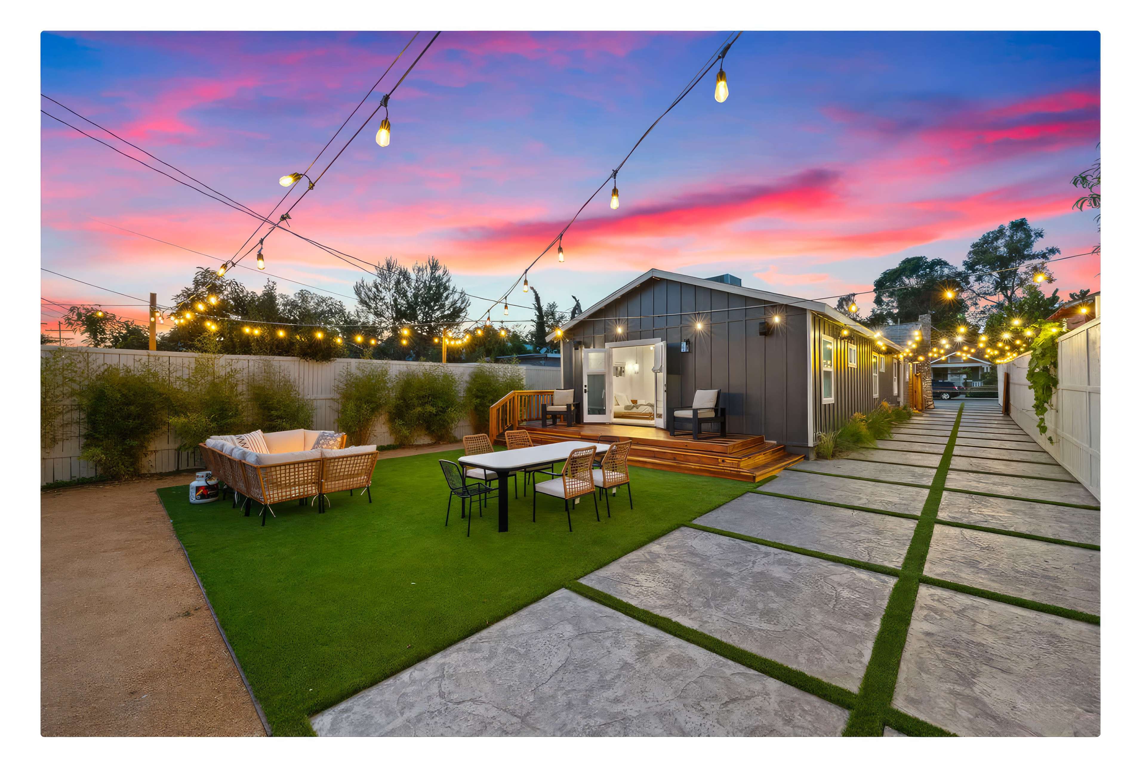 A modern backyard features a patio area with dining furniture, string lights overhead, and a pathway leading to a contemporary house against a vibrant sunset sky.