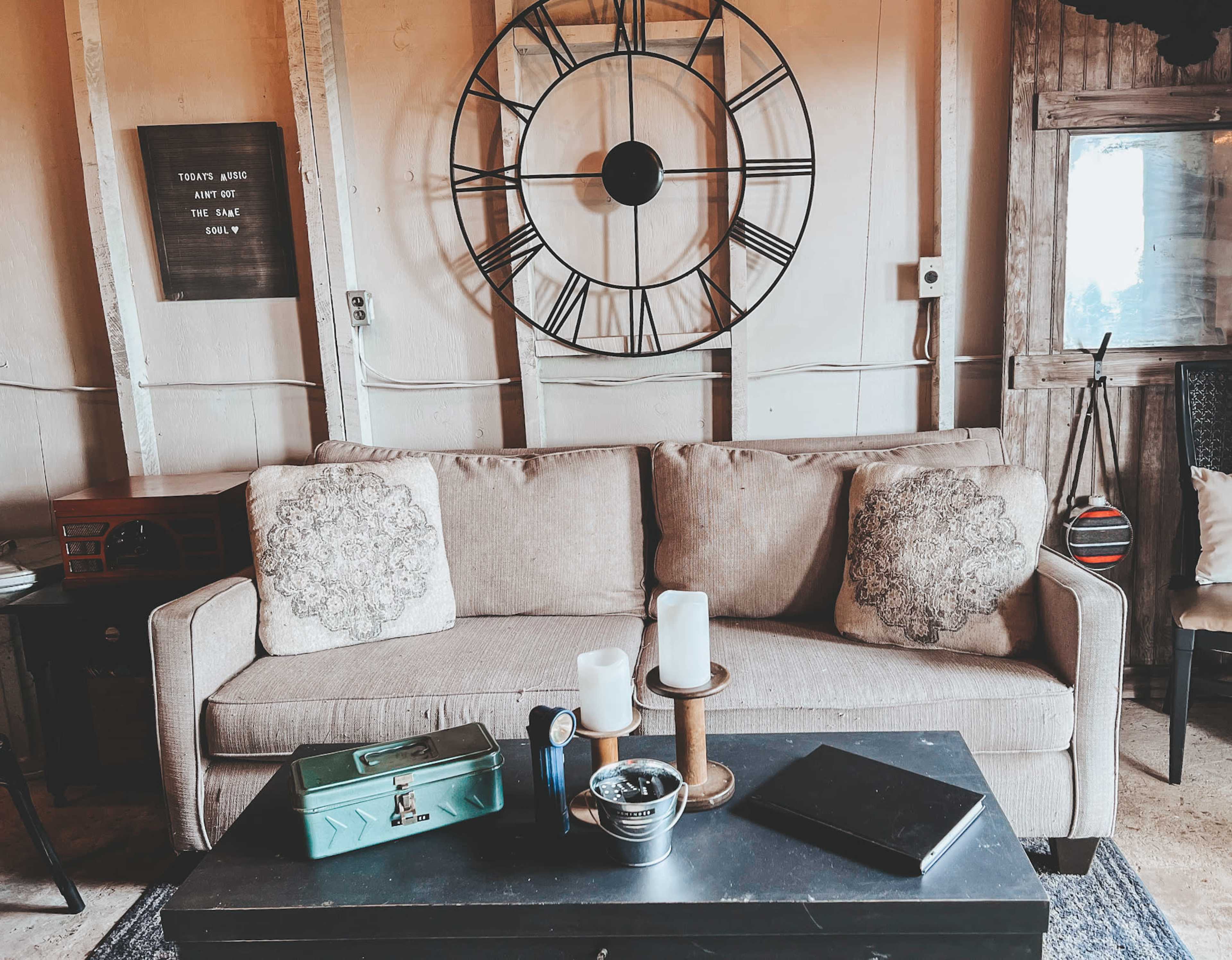 The image shows a living room with a beige sofa adorned with two decorative pillows, a large wall clock, and a black coffee table featuring candles and various items.