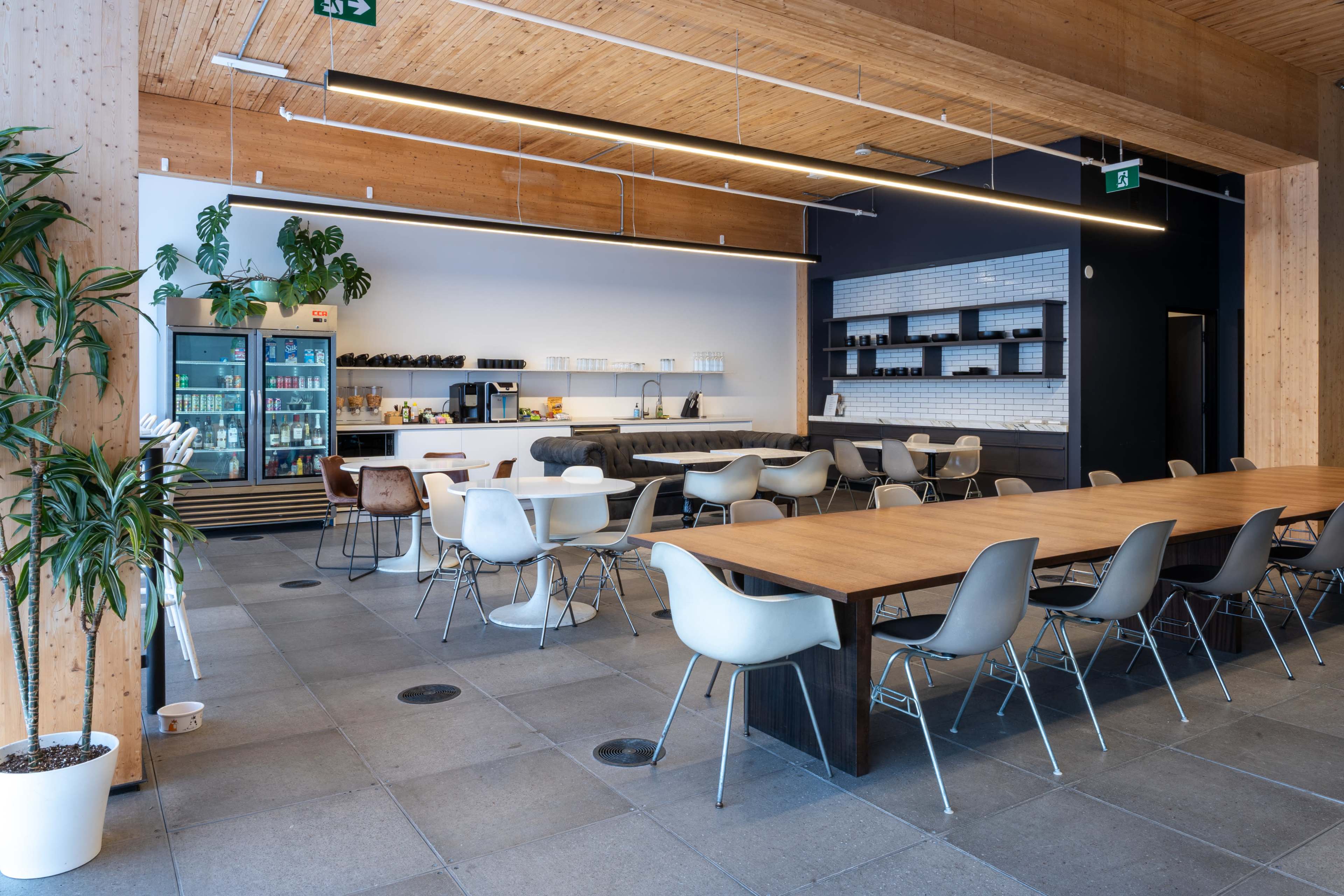 The image shows a modern cafeteria with wooden architecture, a long table, and a beverage refrigerator against a minimalist wall.