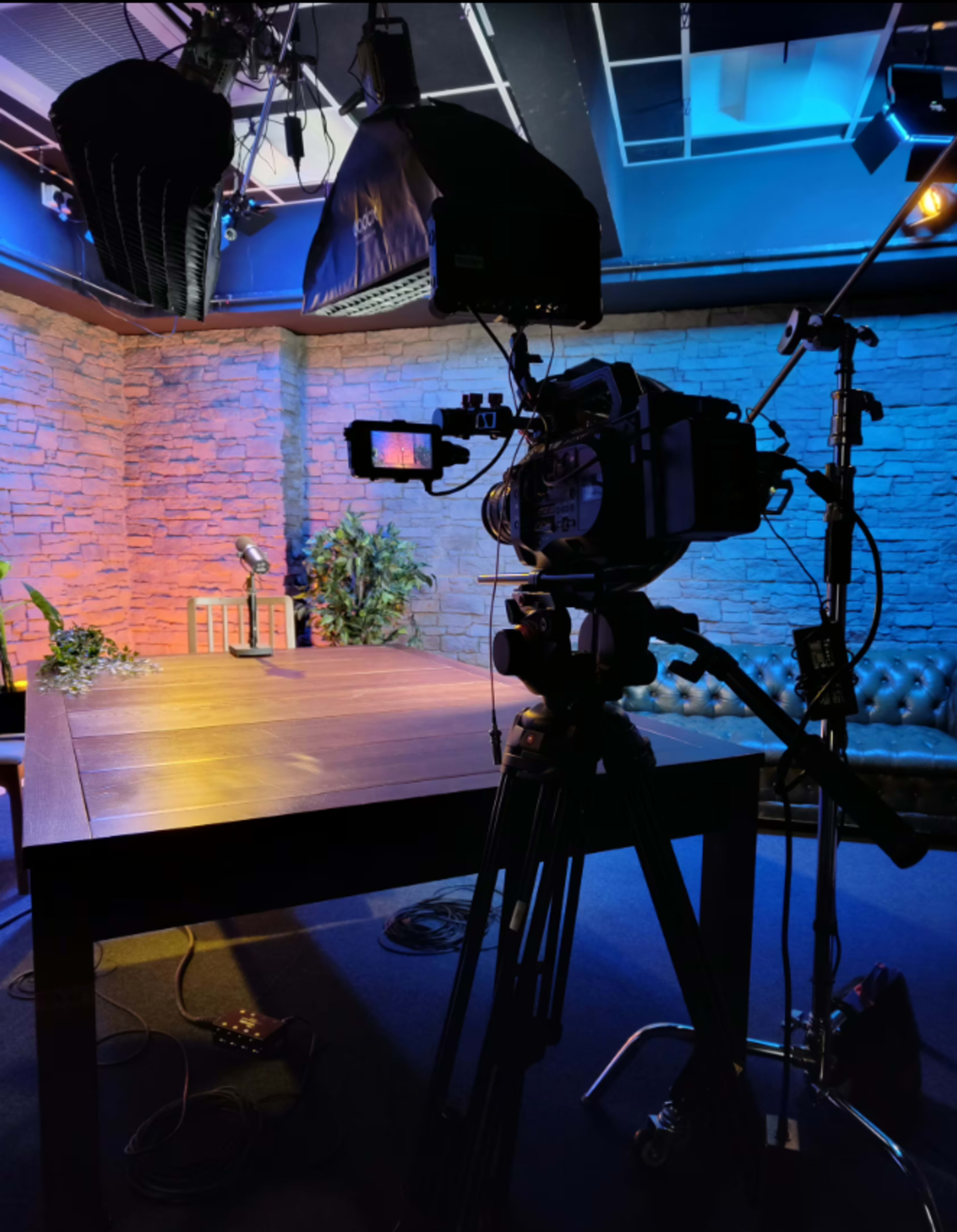 A camera on a tripod is positioned in front of a brightly lit studio set featuring a table and decorative plants against a stone wall backdrop.