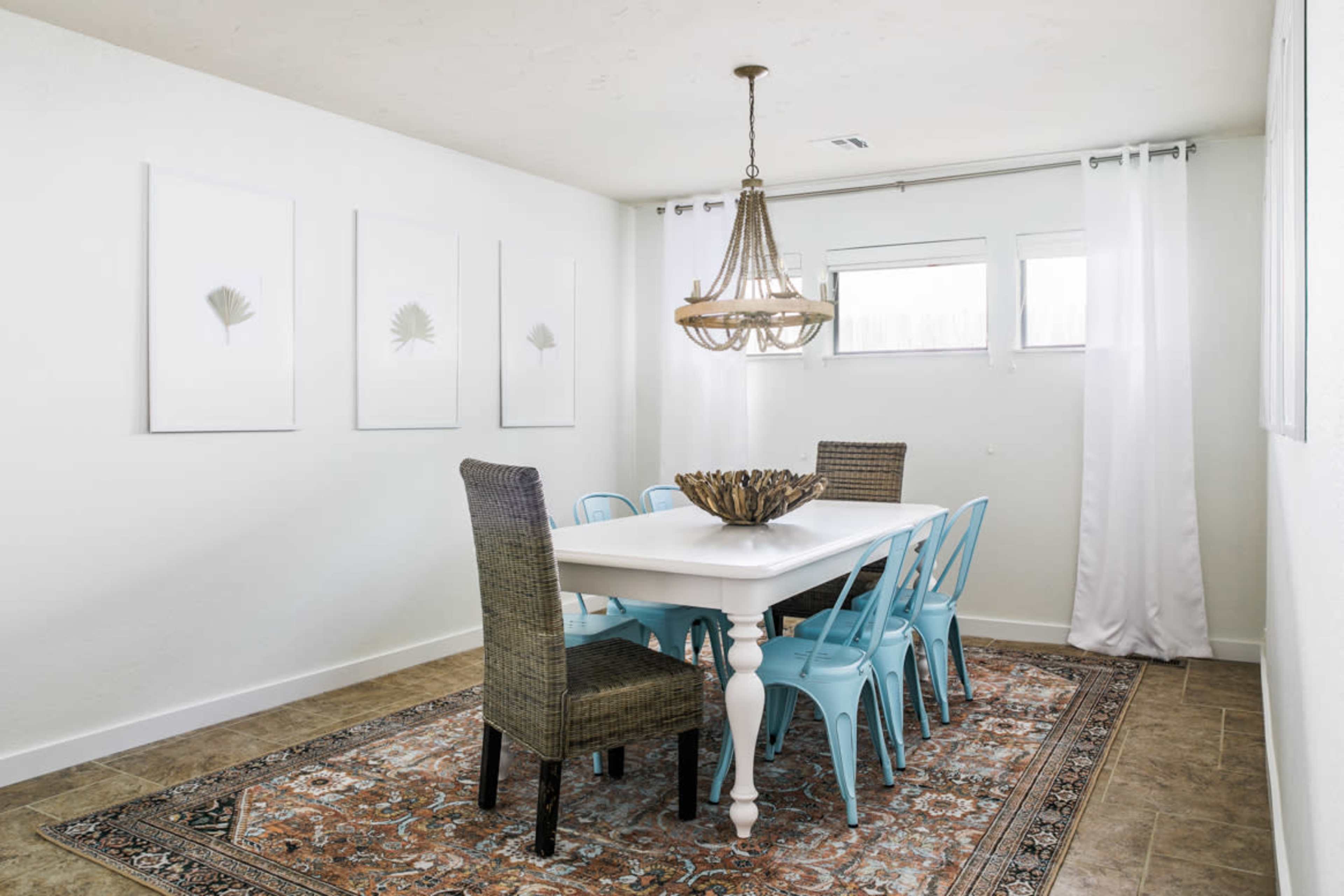 A modern dining room features a long white table surrounded by blue and brown chairs, with a chandelier overhead and framed plant prints on the wall.