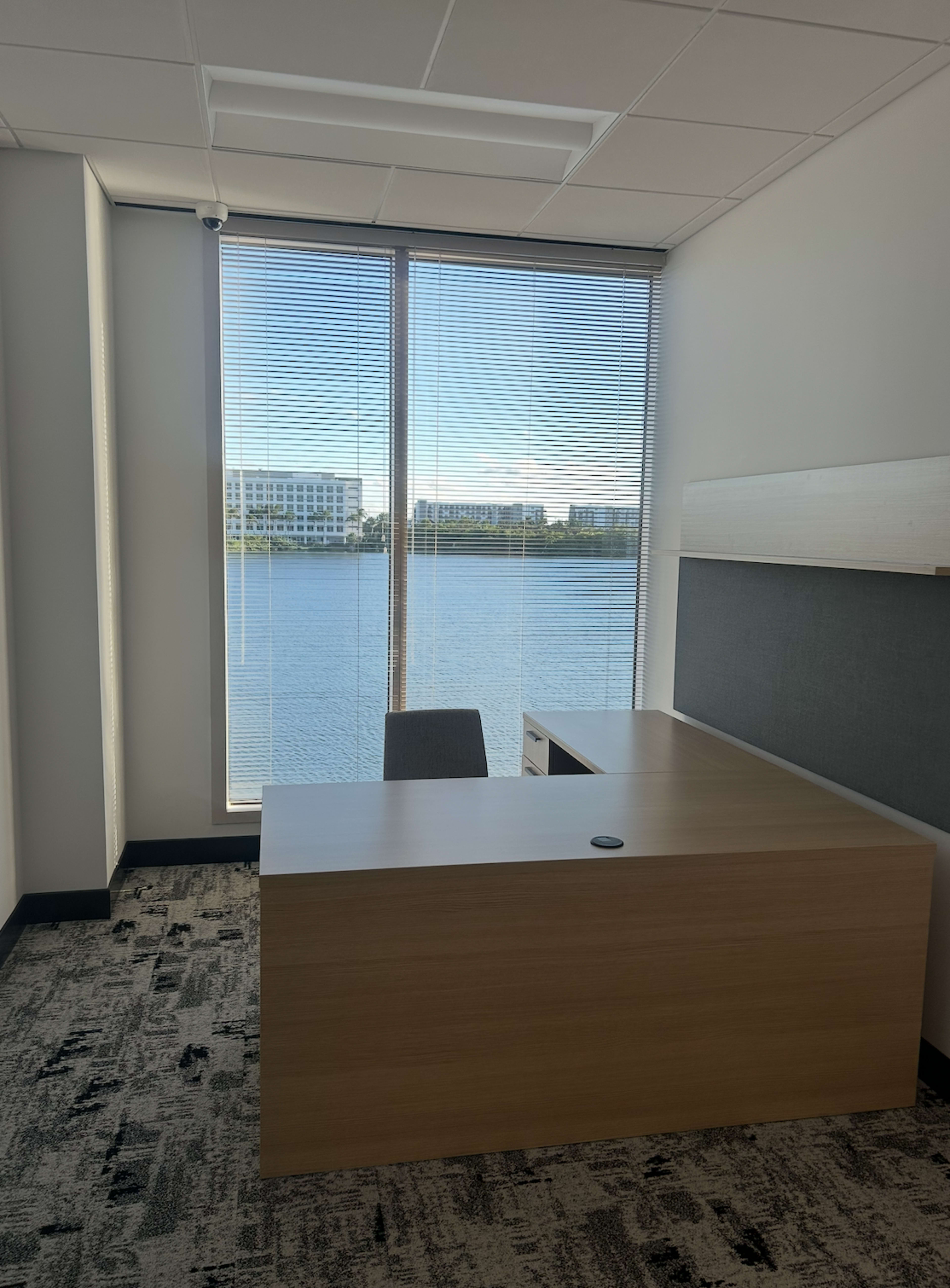 The image depicts a modern office with a wooden desk and chair, overlooking a body of water through a large window.