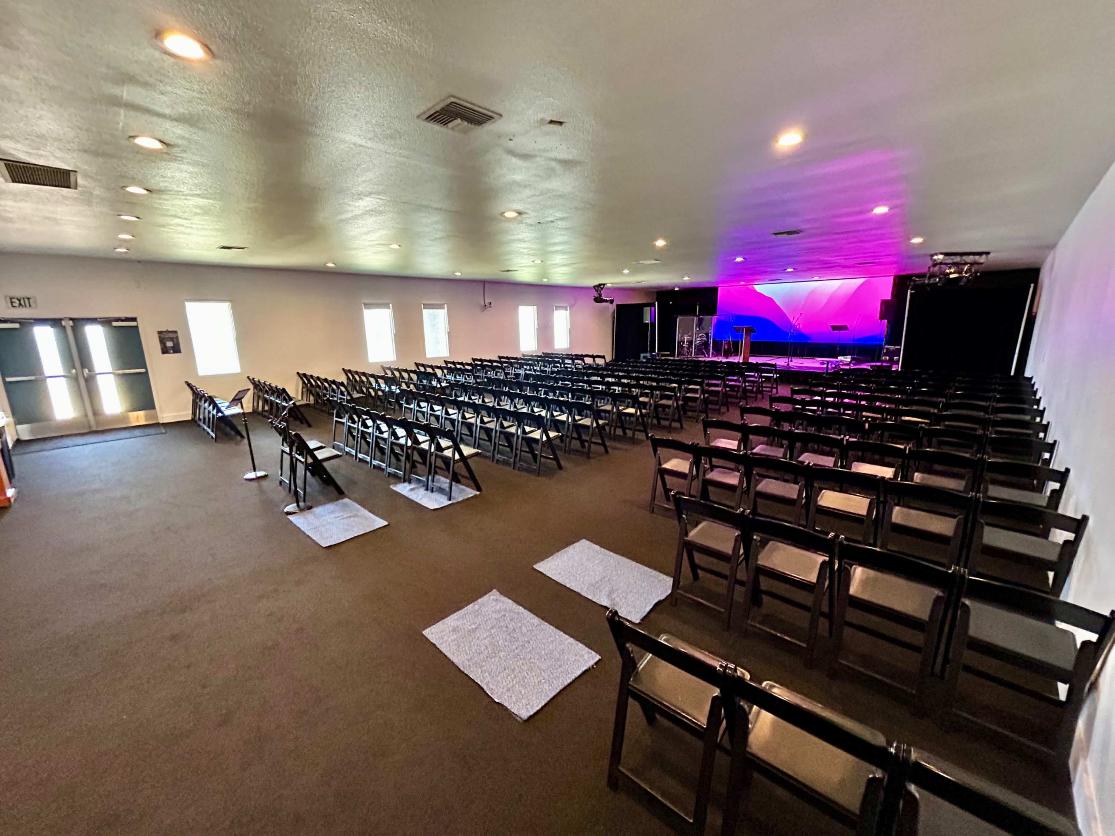 The image shows a spacious indoor venue set up with rows of black folding chairs facing a stage illuminated by colorful lighting.