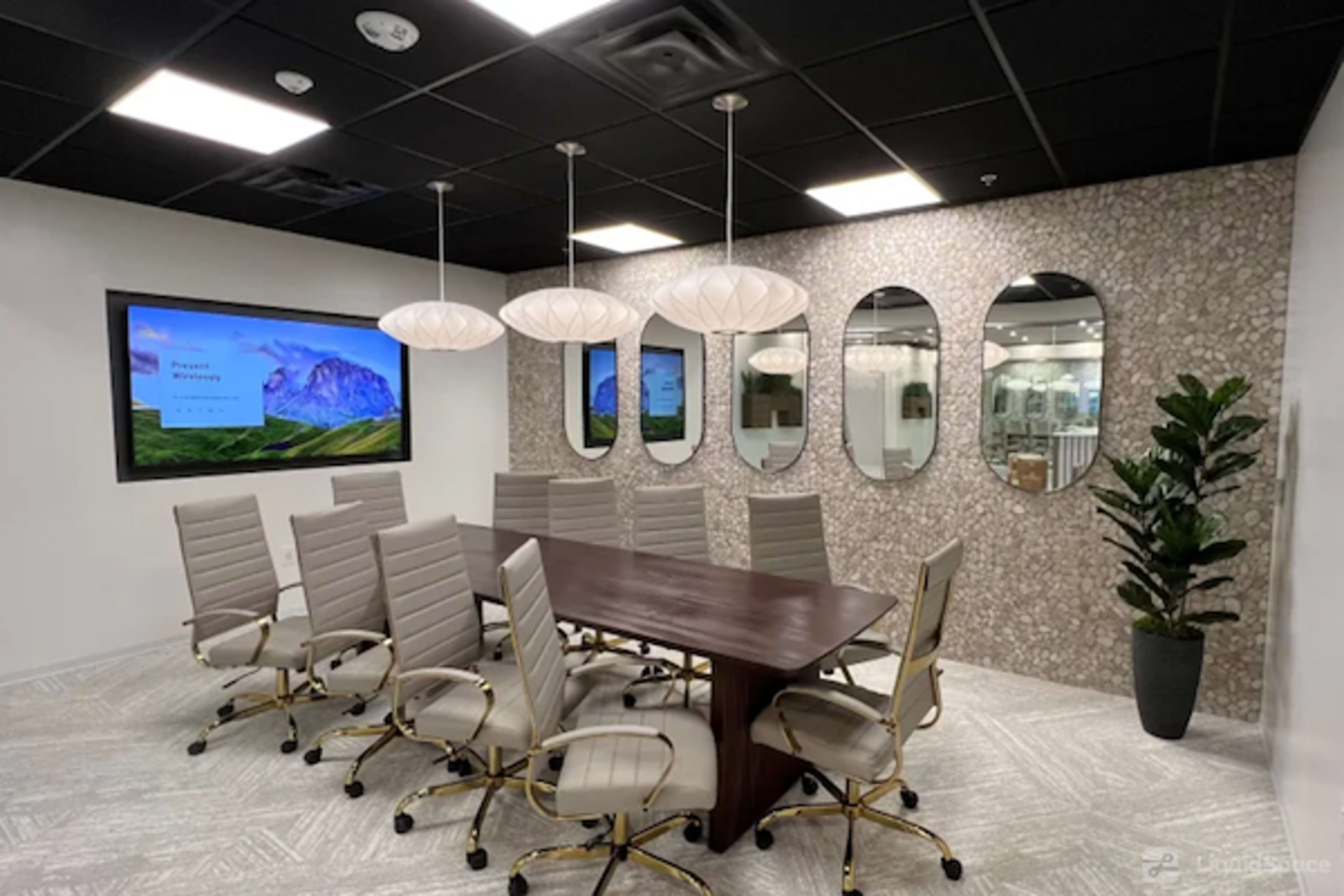 The image shows a modern conference room with a long wooden table, ergonomic chairs, and decorative mirrors on the walls, along with a large screen displaying a presentation.