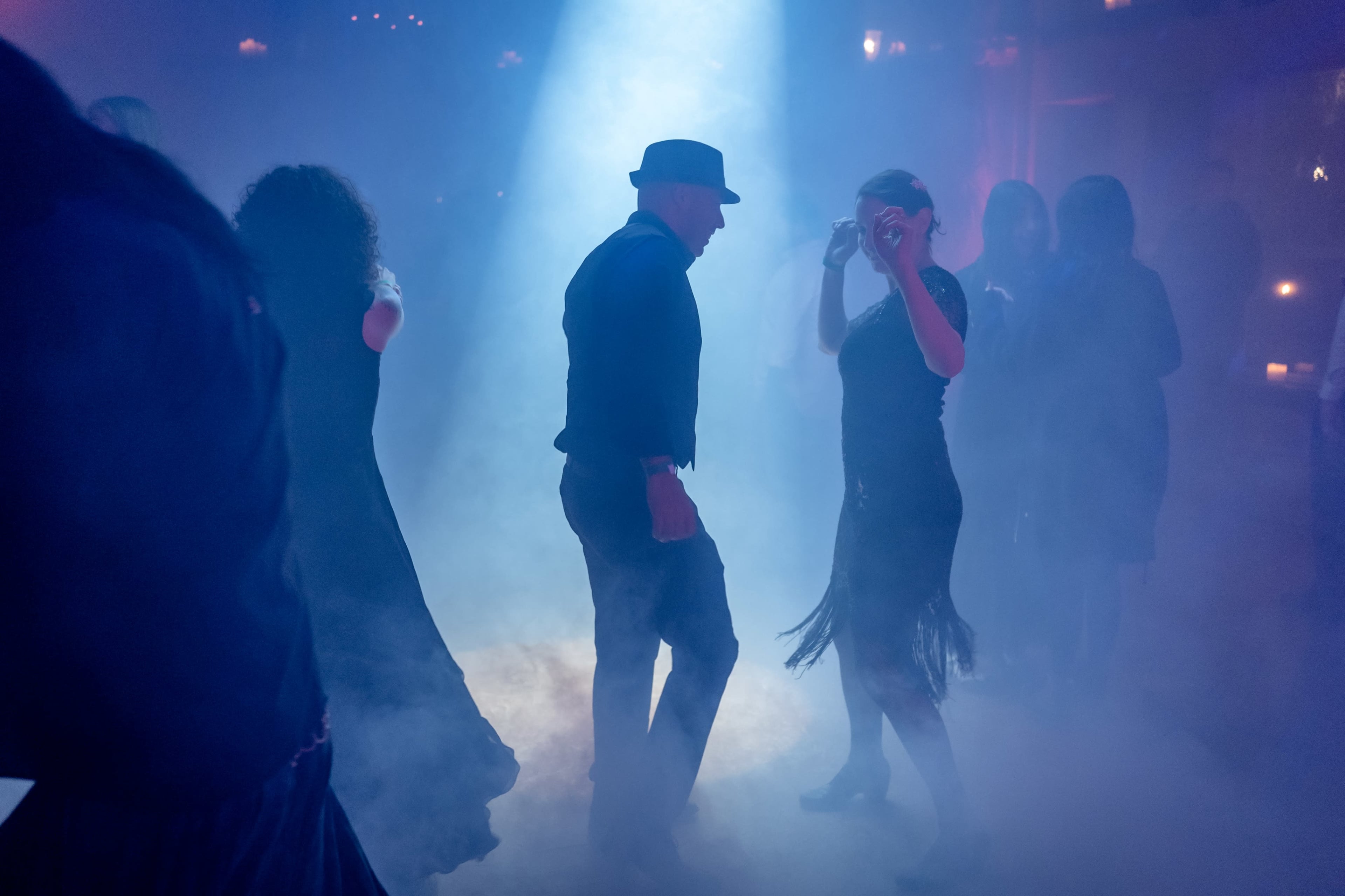 A group of dancers moves in a dimly lit space filled with smoke, highlighted by a beam of light.