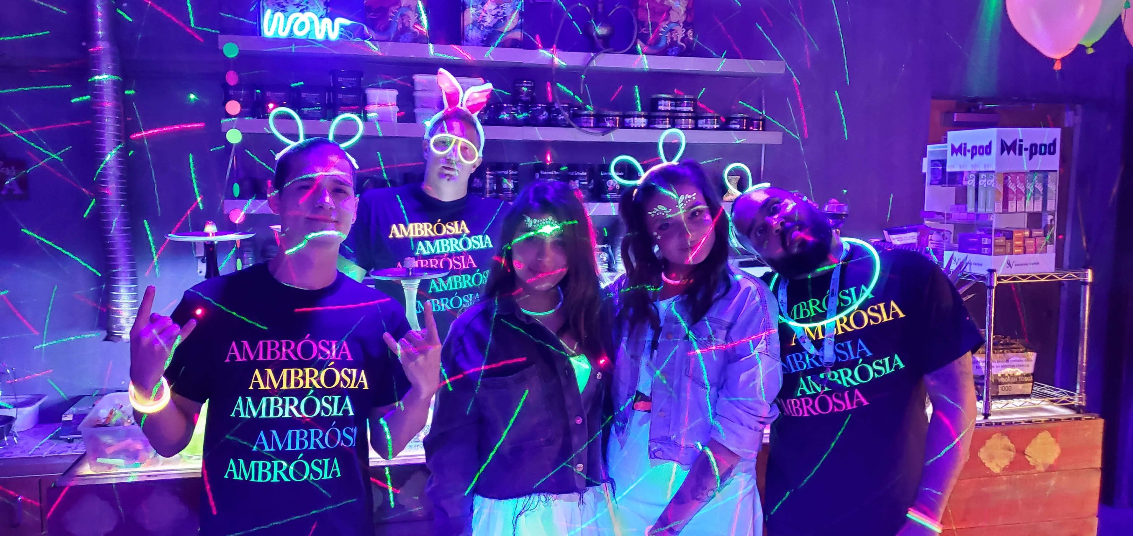 A group of five people wearing matching black shirts with the word "AMBRÓSIA" pose in a room decorated with neon lights and colorful laser effects.