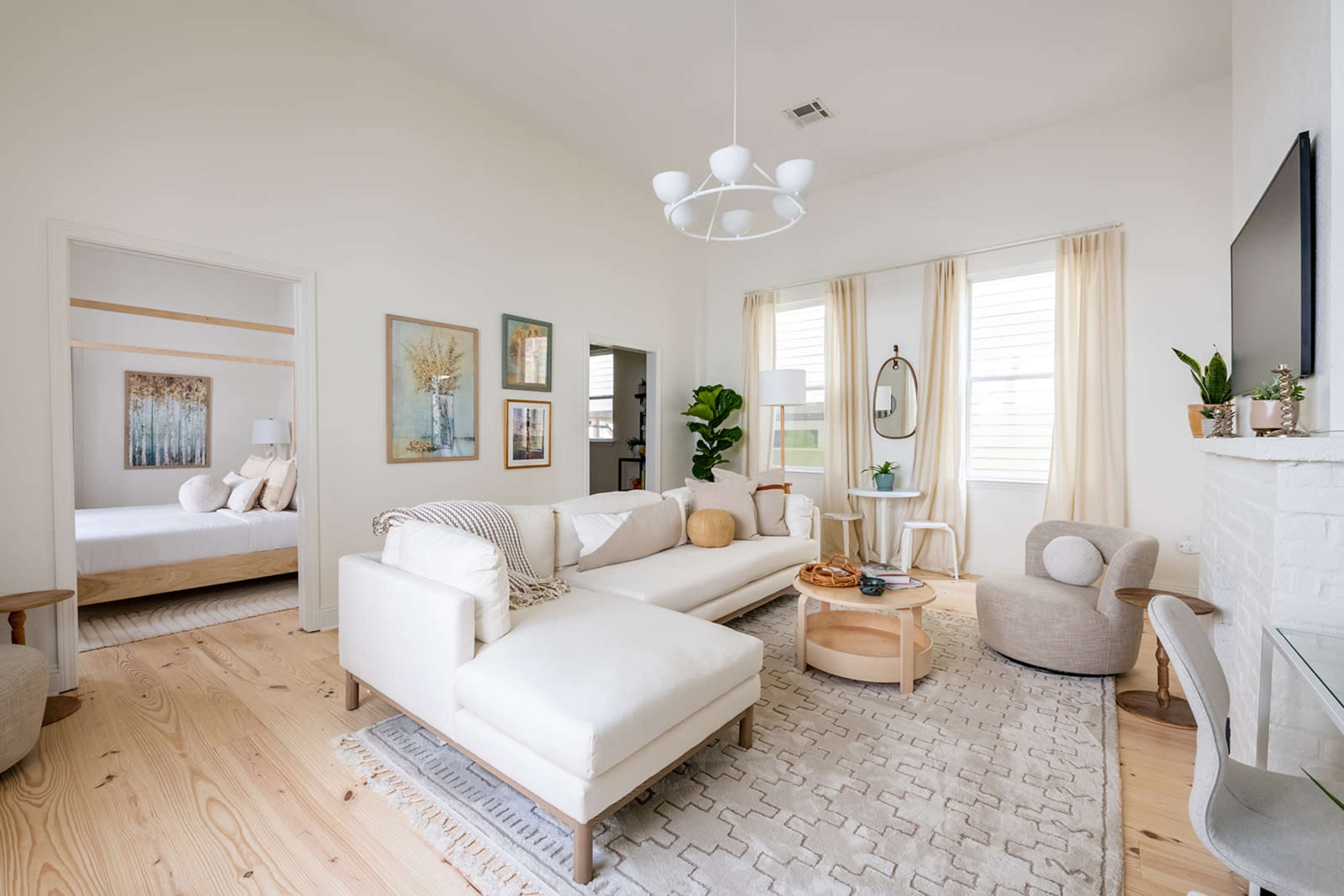 A spacious living room with a light color palette, featuring a white sectional sofa, a round coffee table, and large windows with sheer curtains.