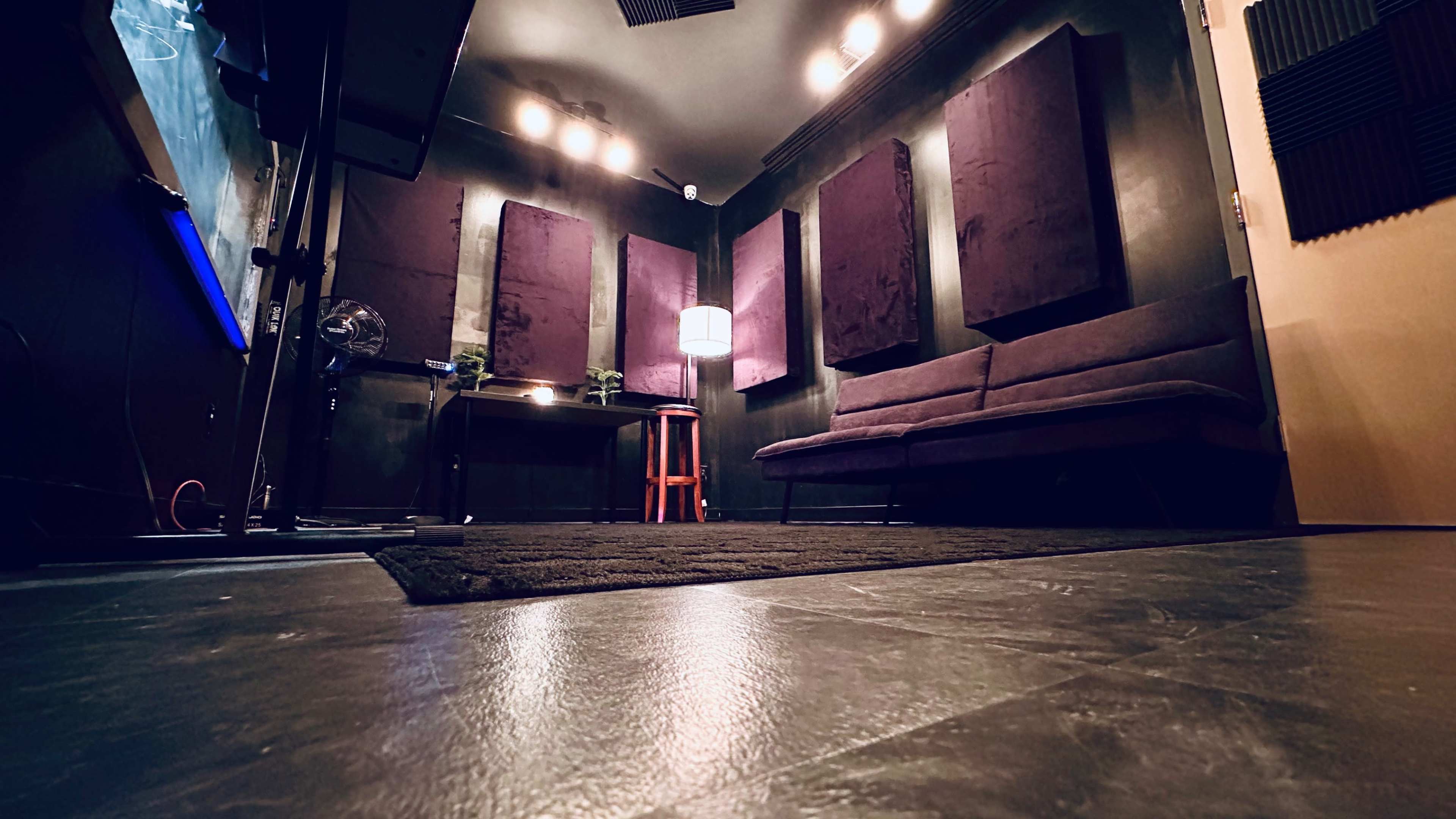 A dimly lit recording studio with purple soundproofing panels on the walls, a table with a lamp, and a couch in the center.