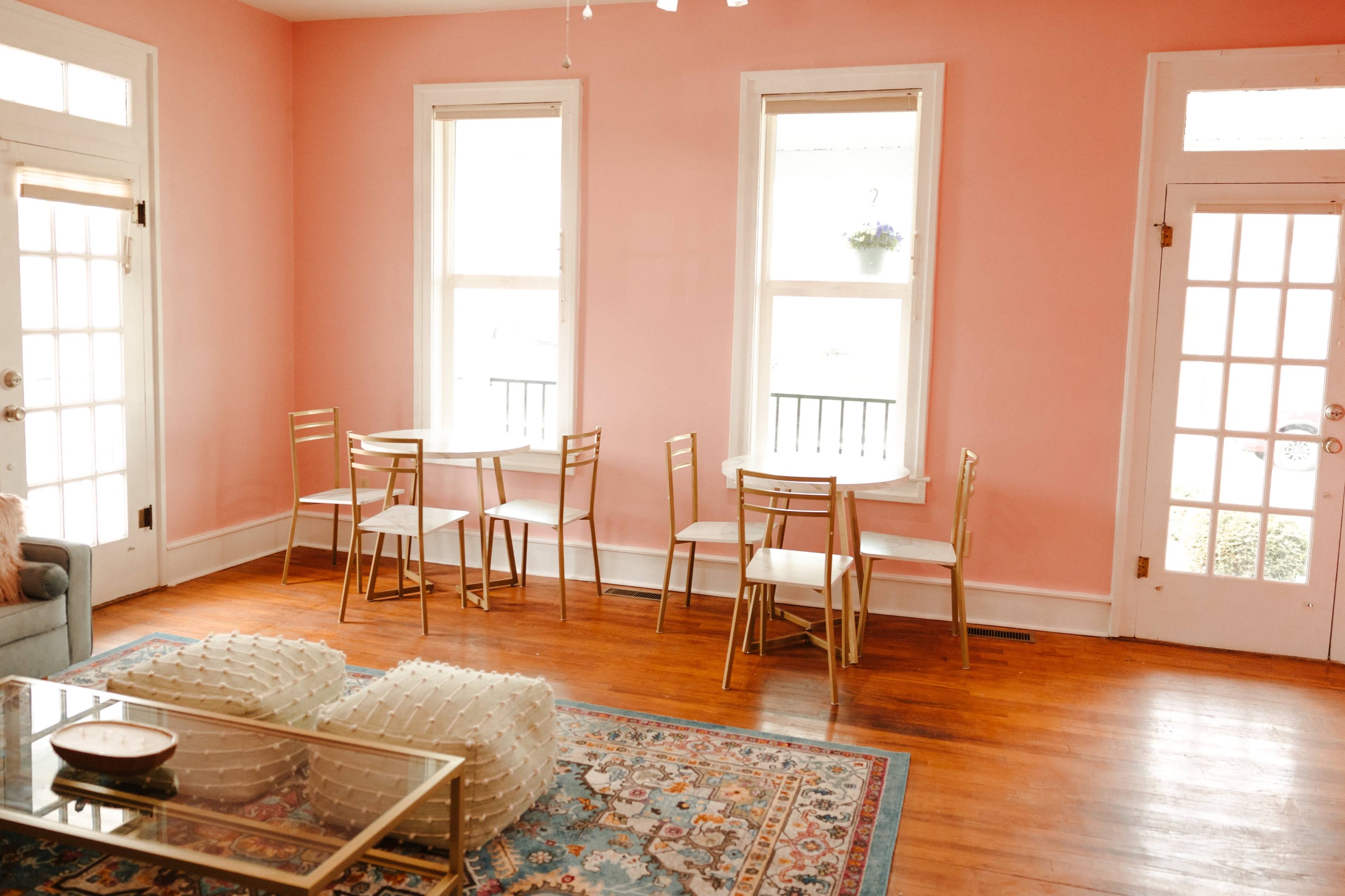 A bright room with pink walls, two large windows, and several gold chairs arranged around small tables.