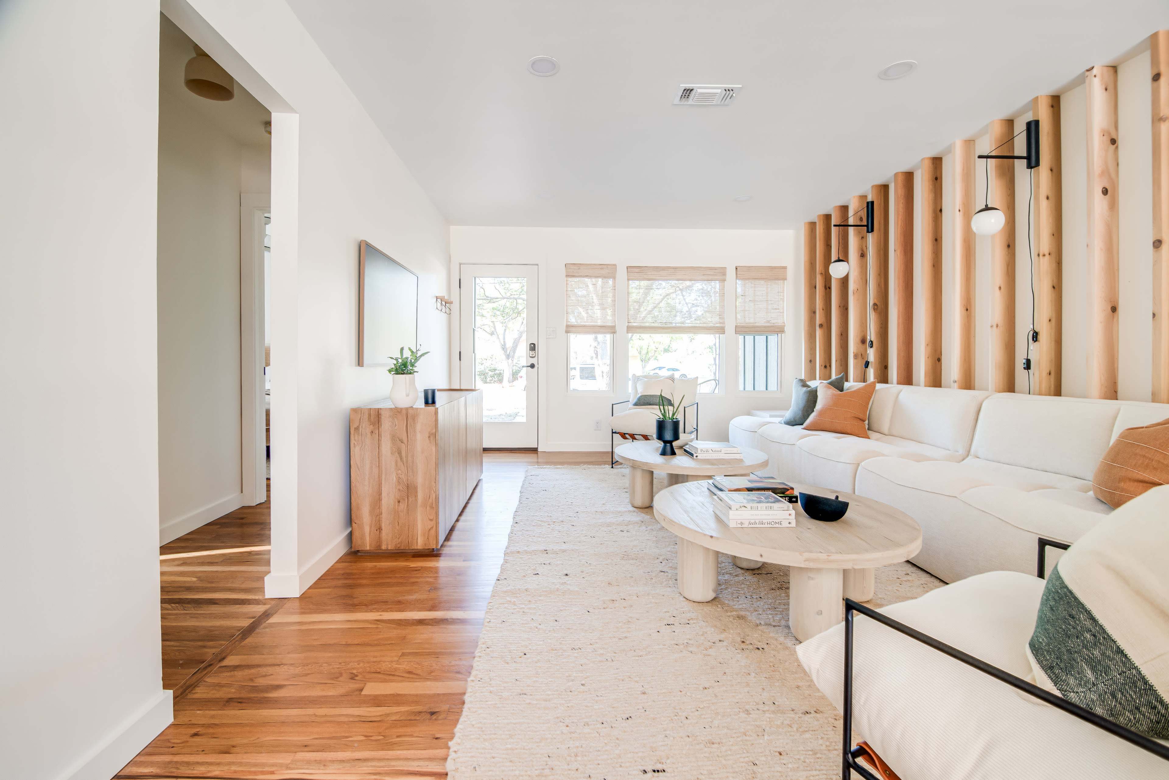 A spacious living room with light-colored walls, a wooden accent wall, a sectional sofa, a round coffee table, and large windows allowing natural light to illuminate the space.