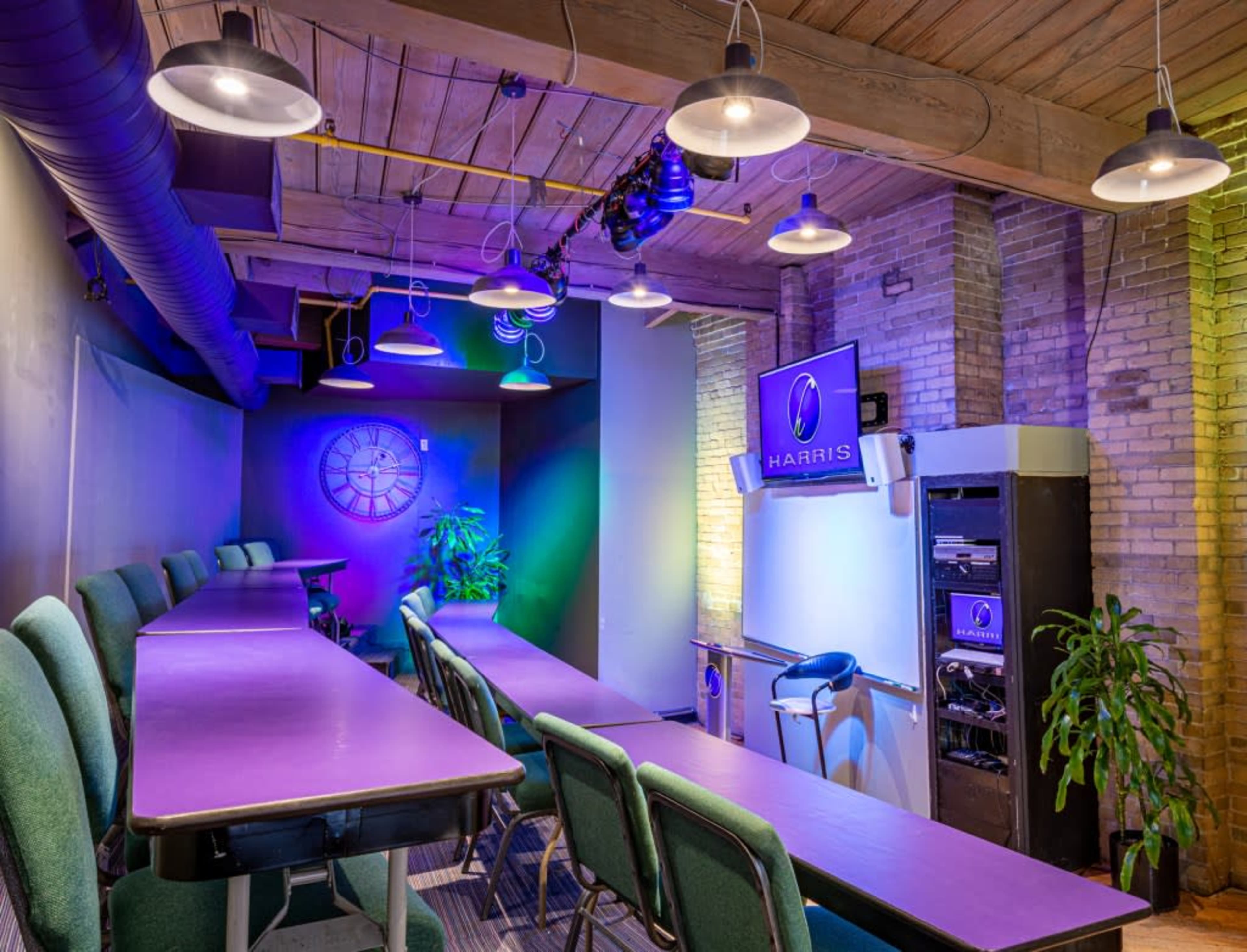 A modern conference room features long purple tables, green chairs, and a wall-mounted screen with the "HARRIS" logo, illuminated by hanging lights.