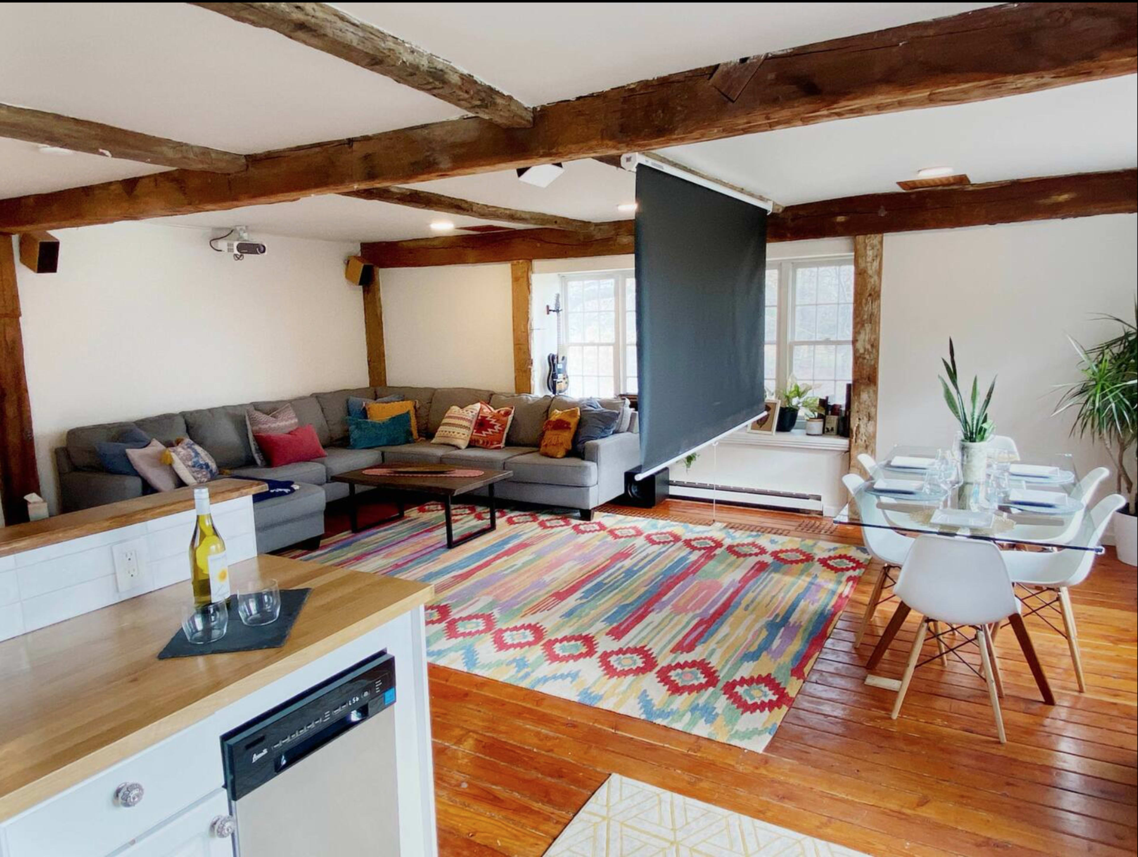 A spacious living area with a gray sectional sofa, a dining table for six, a colorful area rug, and wooden beams on the ceiling.