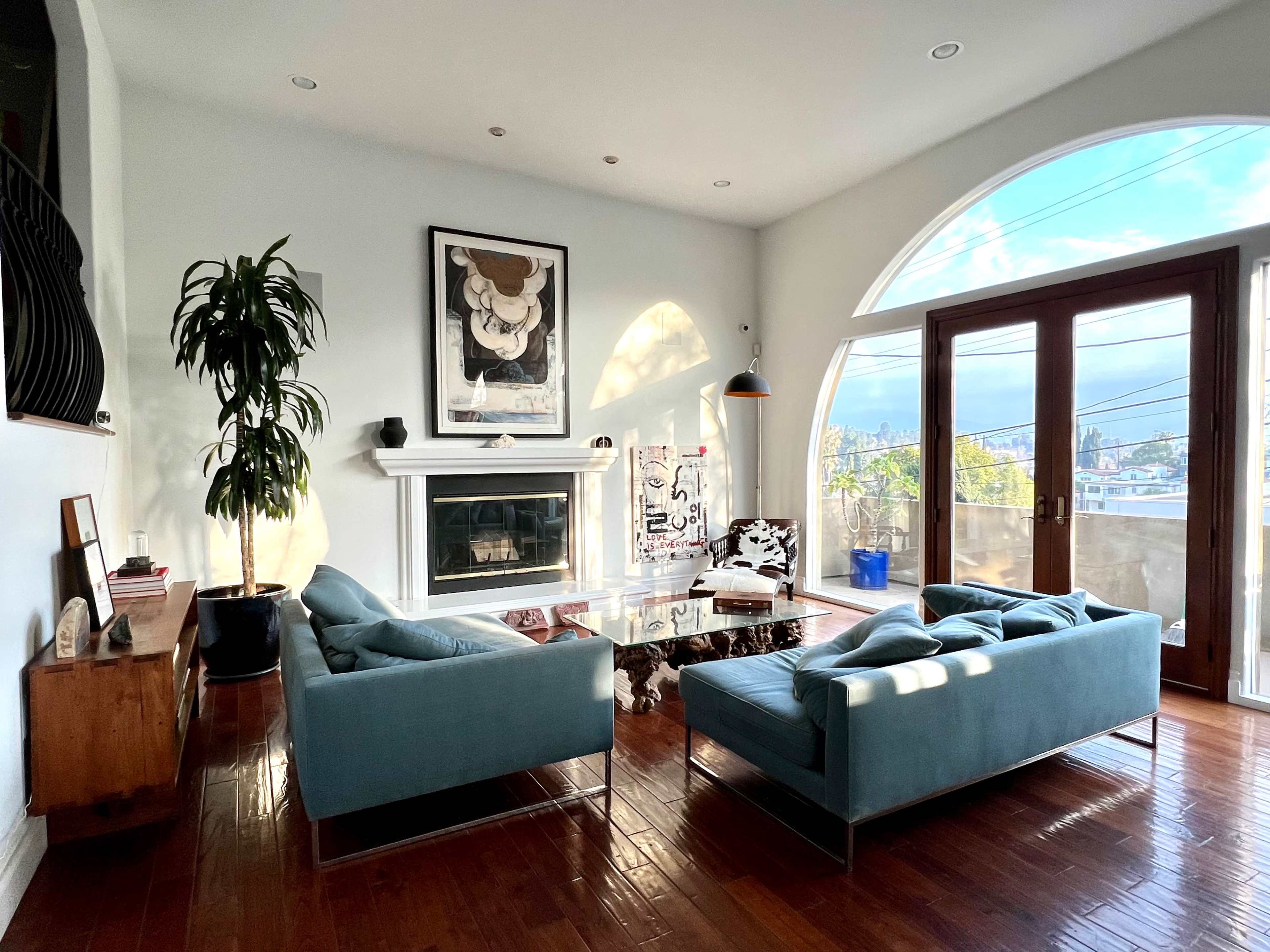 The living room features two blue sofas facing a glass coffee table in front of a white fireplace, with large windows offering a view of the outside.