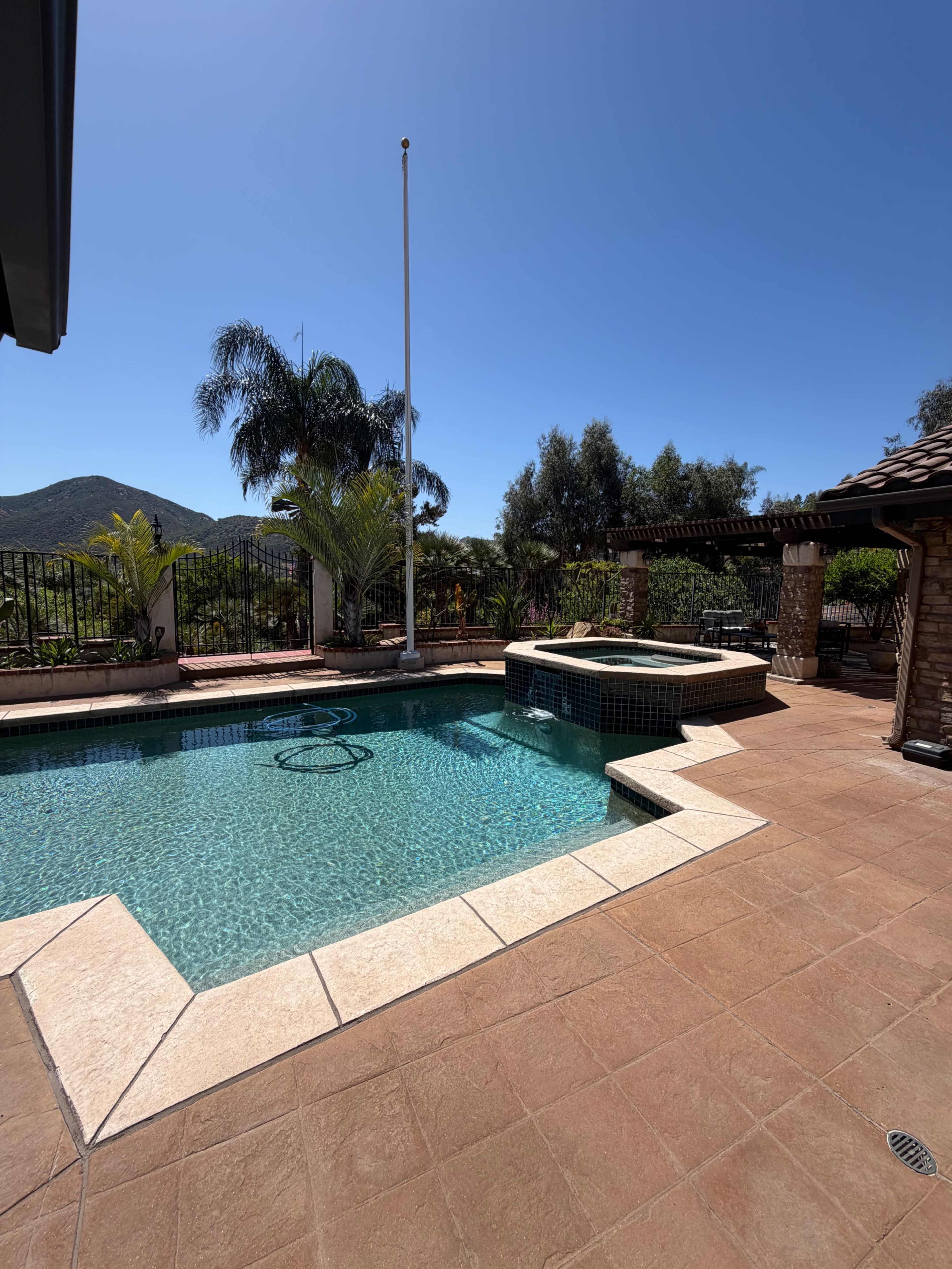The image shows a swimming pool with a spa, surrounded by landscaped greenery and a mountain in the background under a clear blue sky.