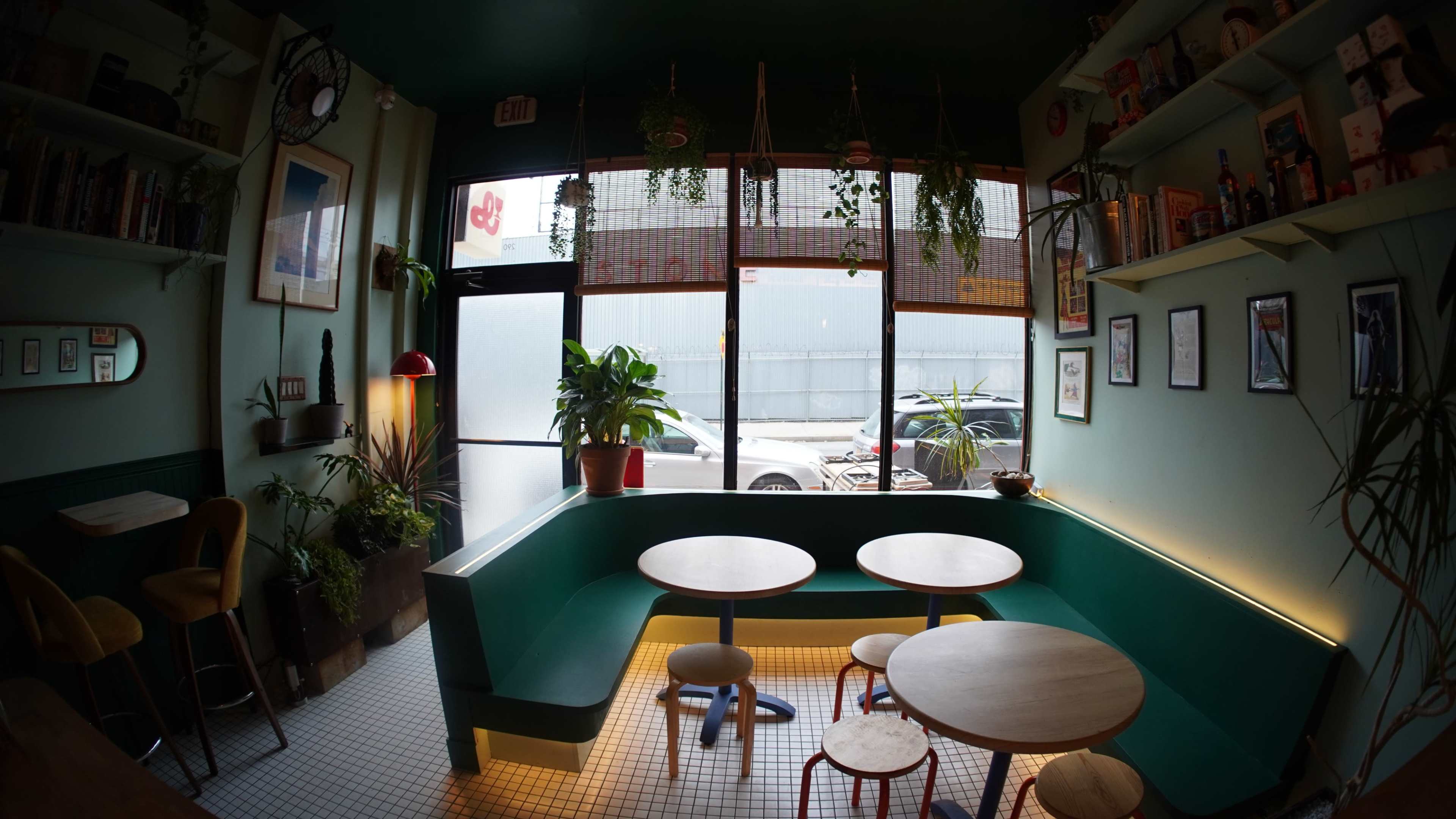 The image shows a cozy, modern café space with green walls, two round tables, and large windows displaying a view of the street outside.