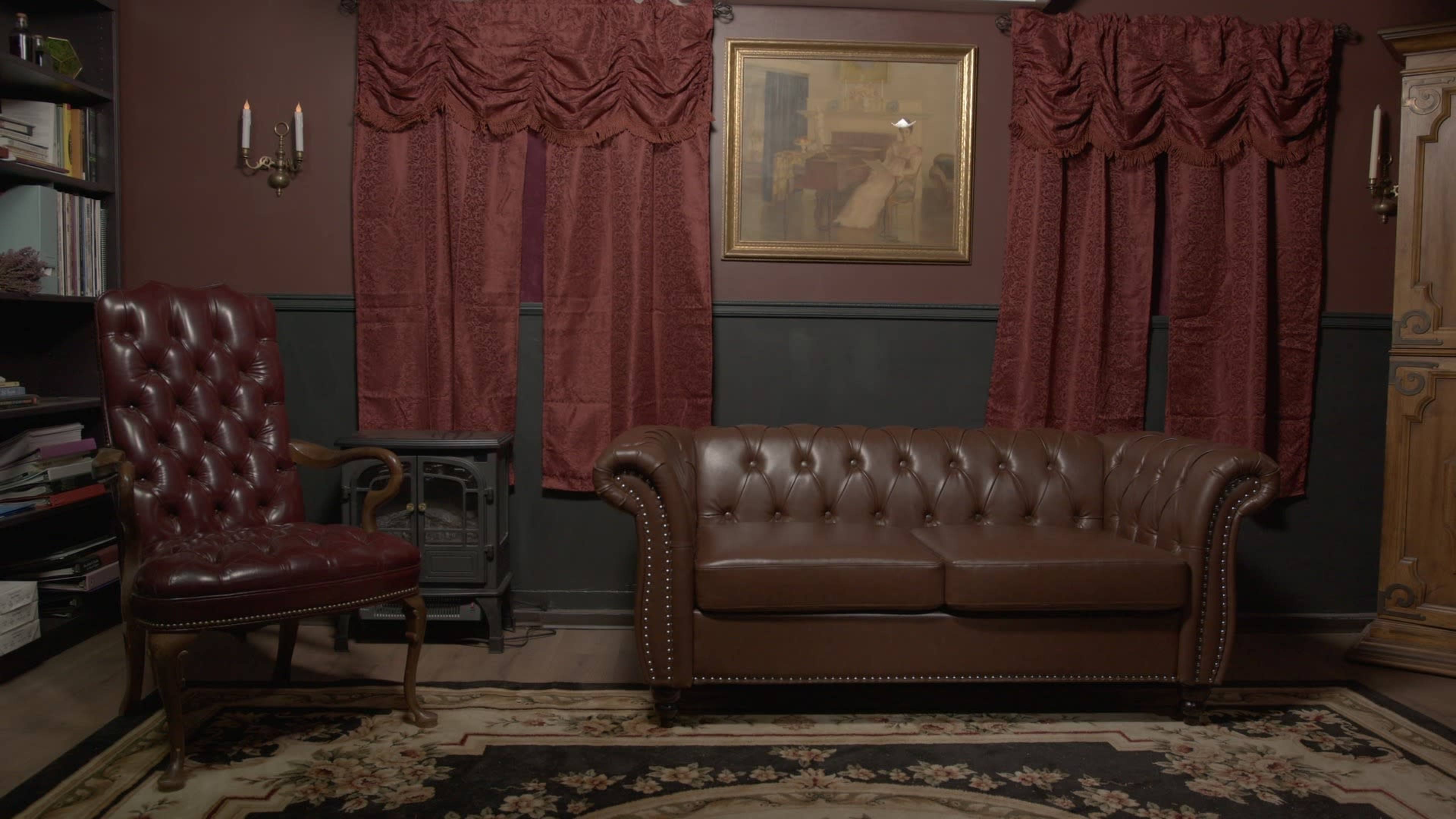The image shows a darkly colored room featuring a brown leather chesterfield sofa, a burgundy wingback chair, dark curtains, and a framed painting on the wall, alongside a small stove.