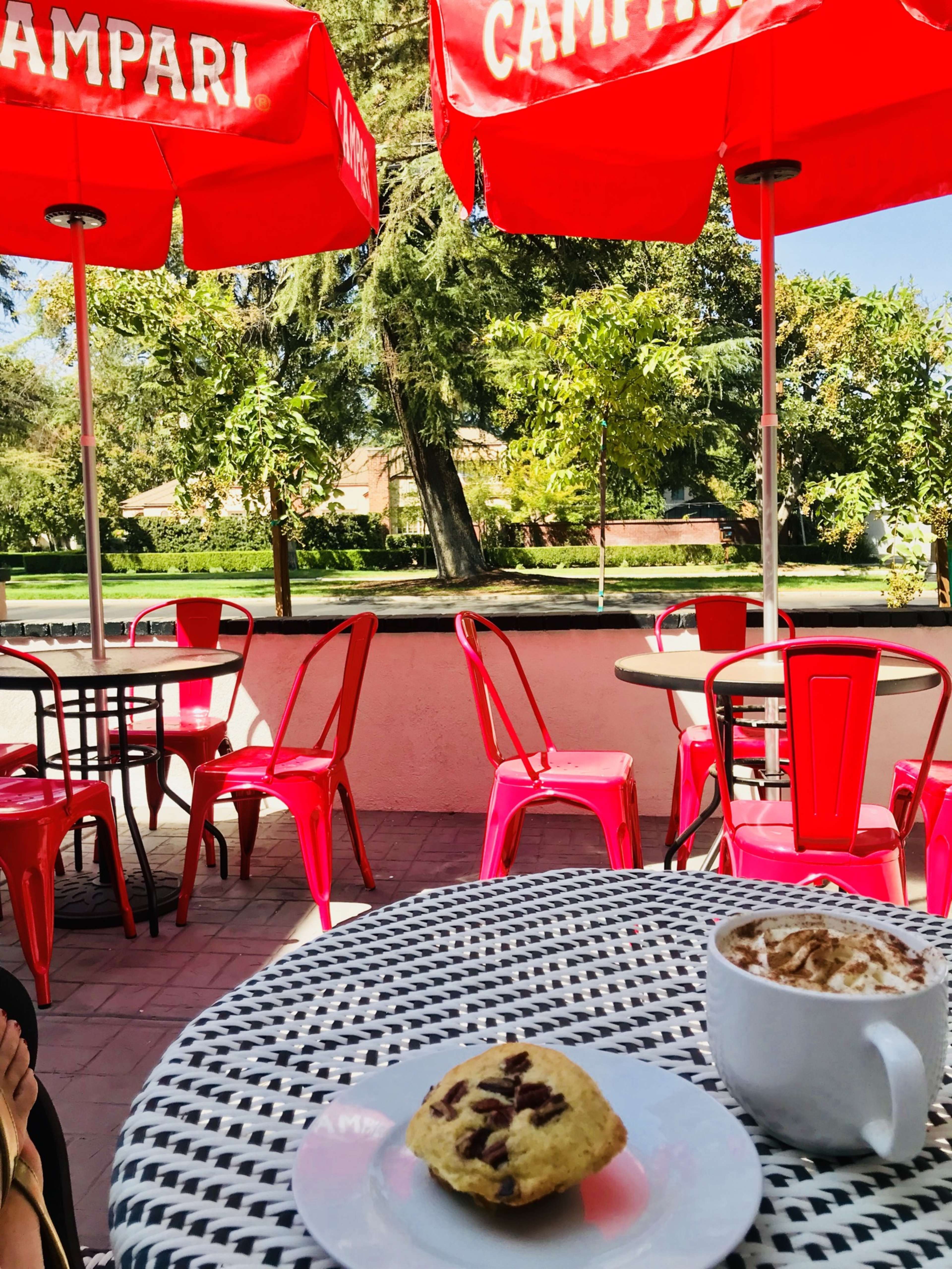 A table with a plate of cookies and a cup of coffee sits in a café with red umbrellas and outdoor seating in a park-like setting.