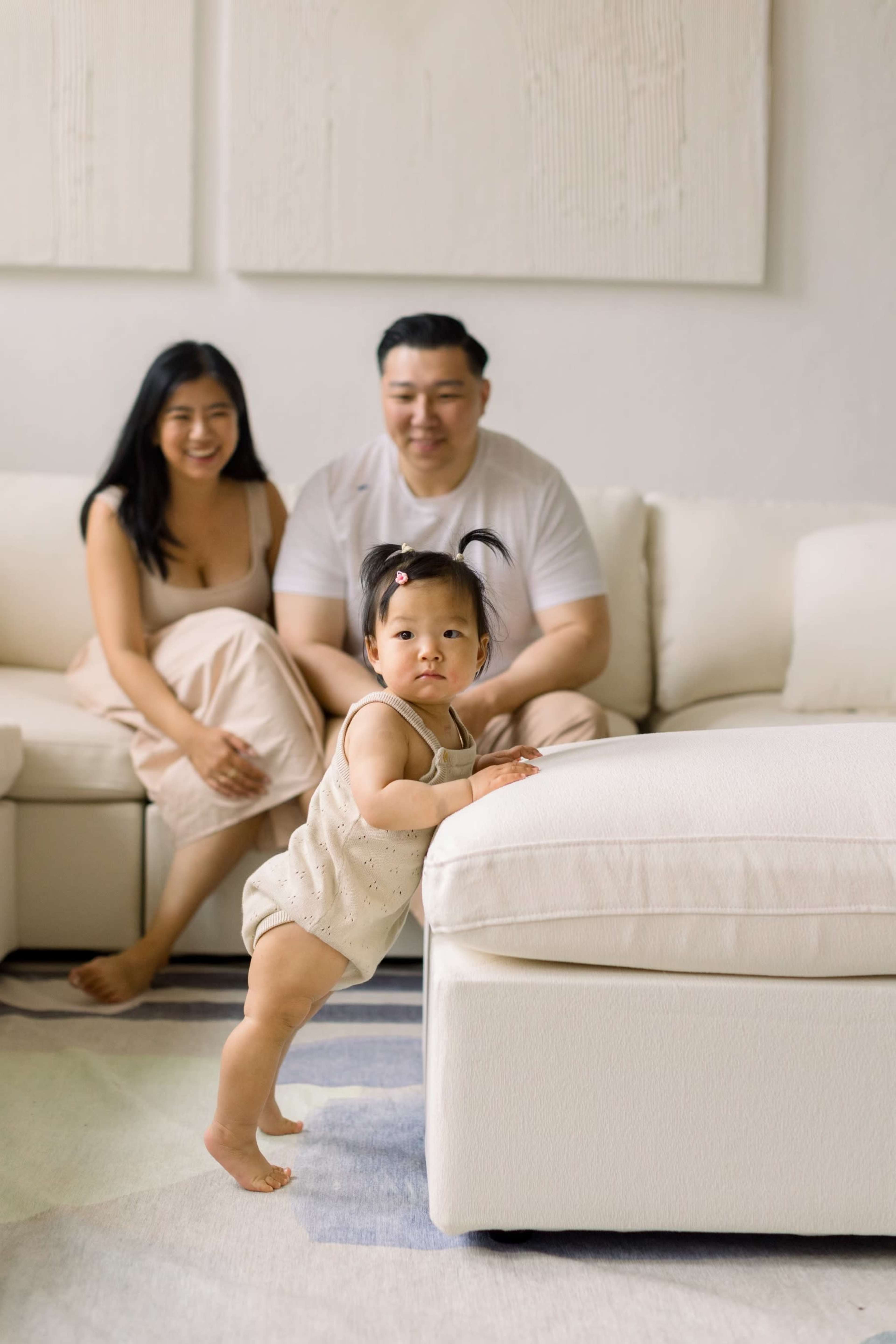 A young child climbs on the edge of a couch while two adults sit on the sofa behind her.