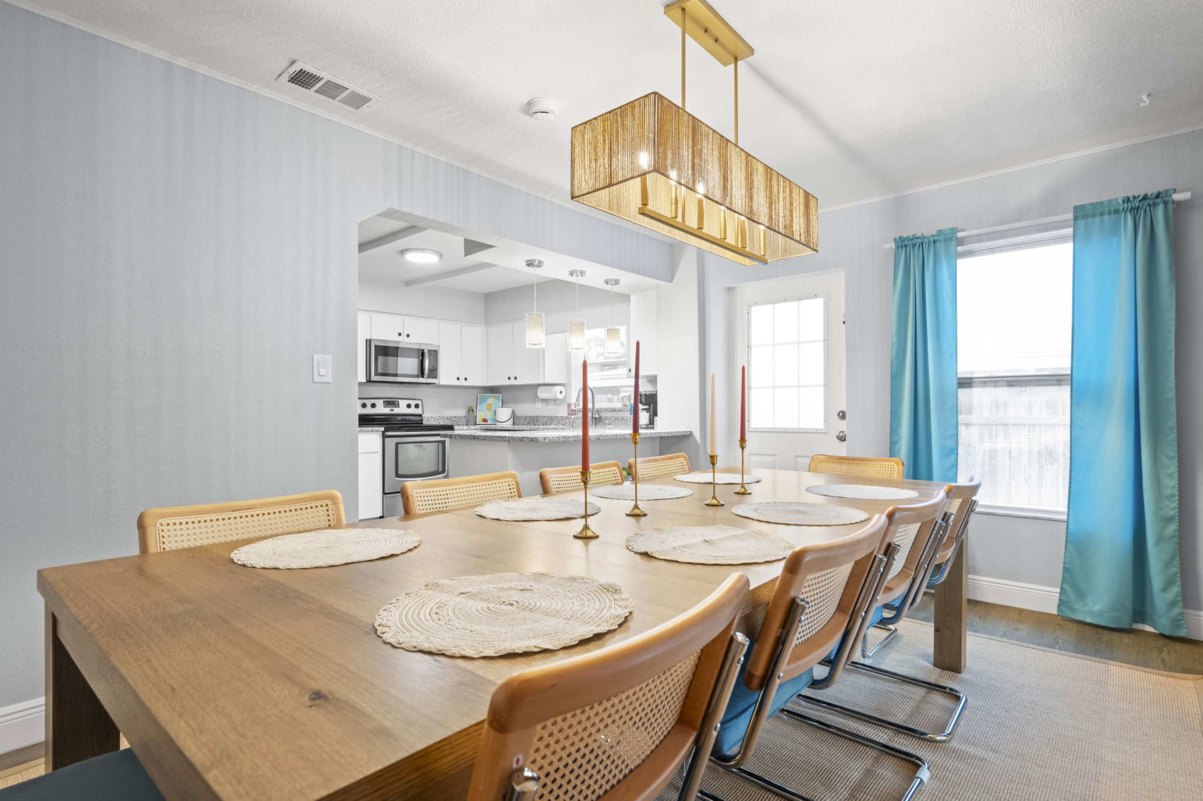 A dining room features a large wooden table with woven placemats and eight chairs, illuminated by a modern pendant light, while turquoise curtains frame a window.