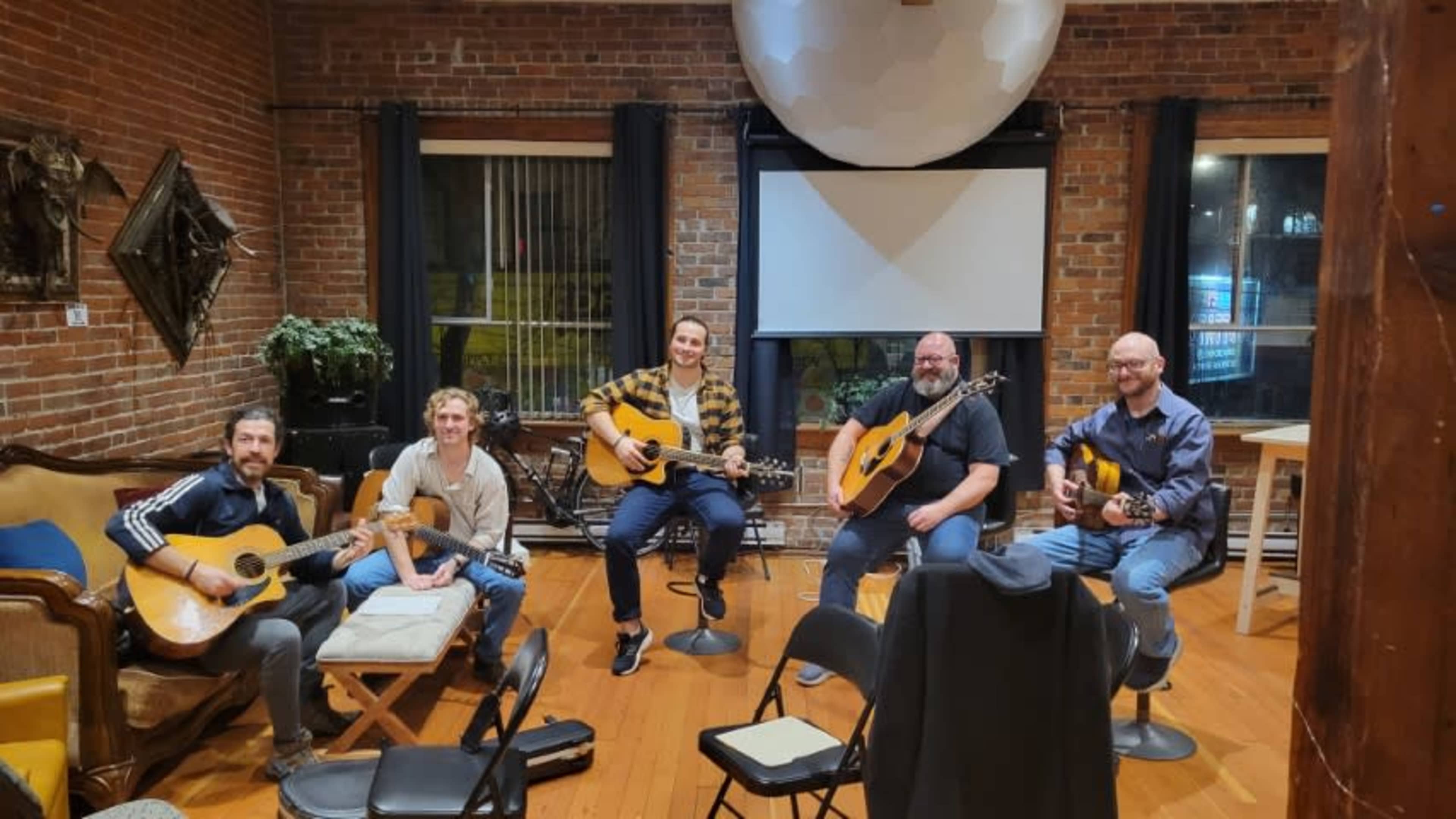 Five musicians sit in a cozy room with brick walls, each holding an acoustic guitar, while a large screen is displayed in the background.