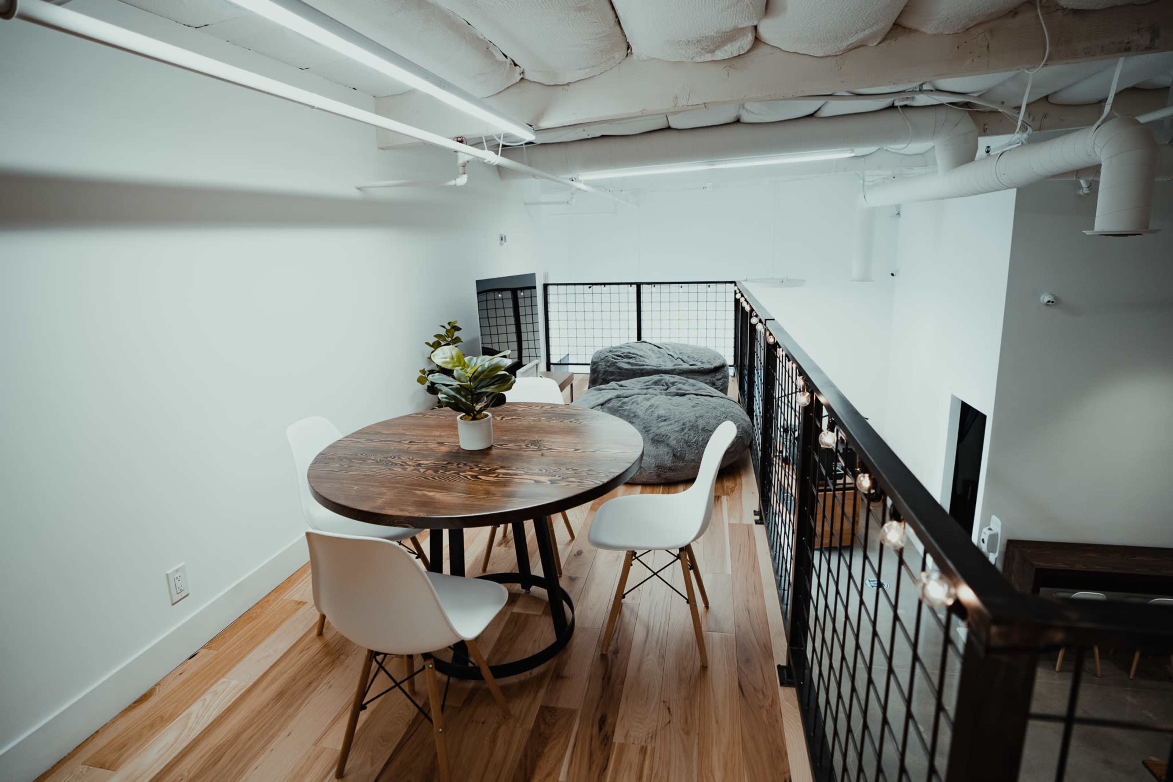 A circular wooden table with white chairs is positioned in a well-lit loft area overlooking a lower space with large bean bags.