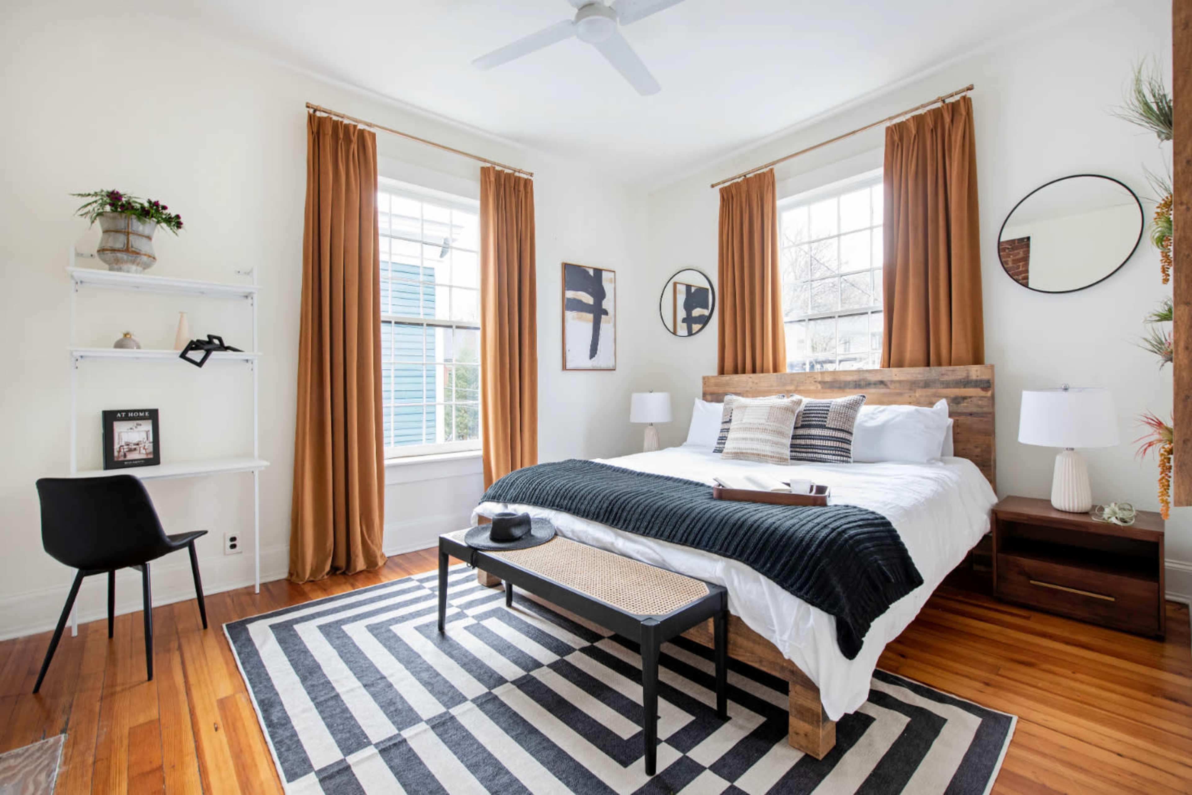 The image shows a bright bedroom featuring a wooden bed frame, a striped area rug, and large windows with cream-colored walls and orange drapes.