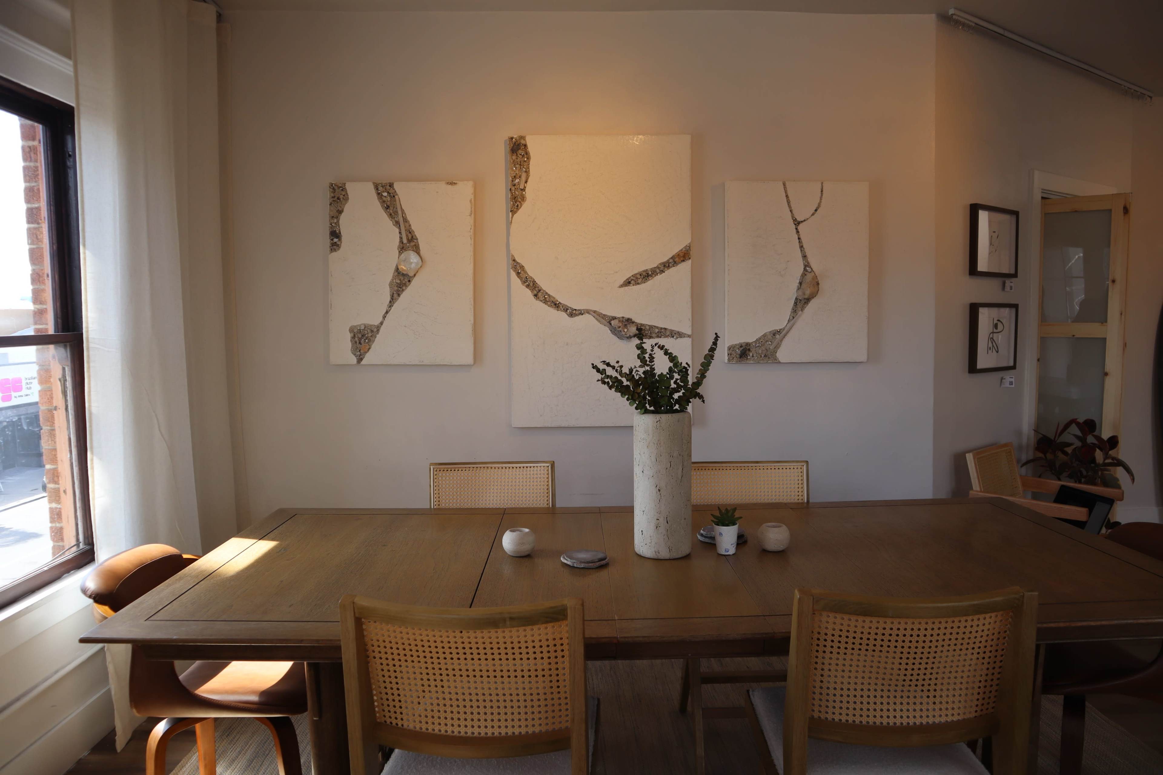 The image shows a dining room featuring a wooden table surrounded by cane-backed chairs, with three abstract artworks hanging on the wall above a tall, cylindrical vase containing greenery.