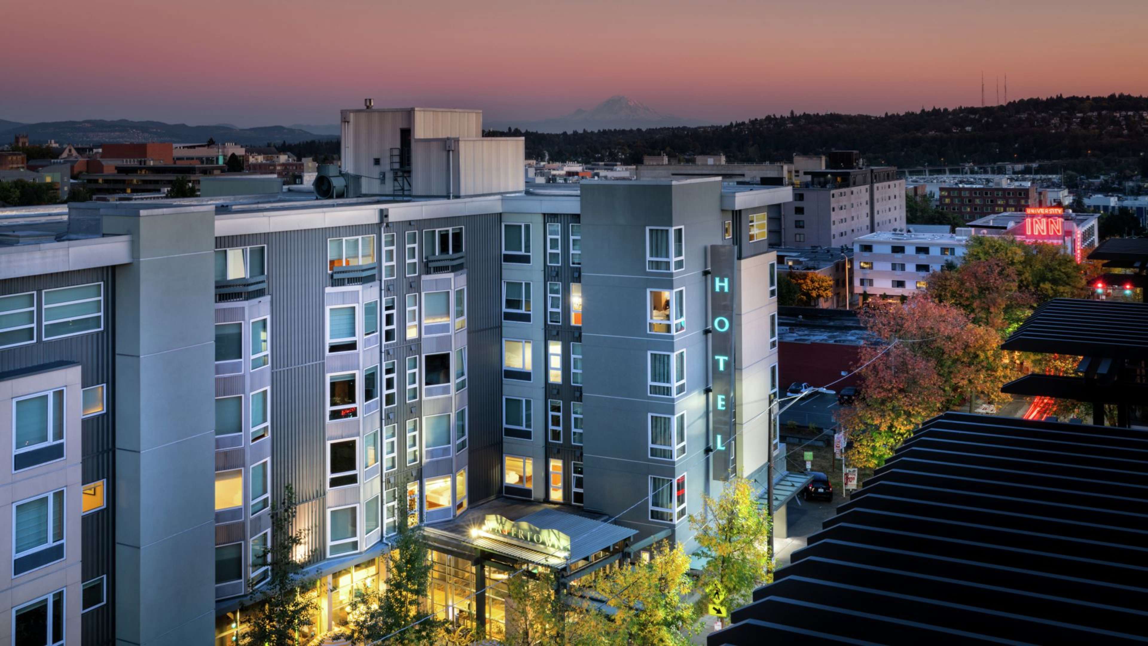 A modern hotel building in an urban setting at dusk, with city lights illuminated and a mountain visible in the background.