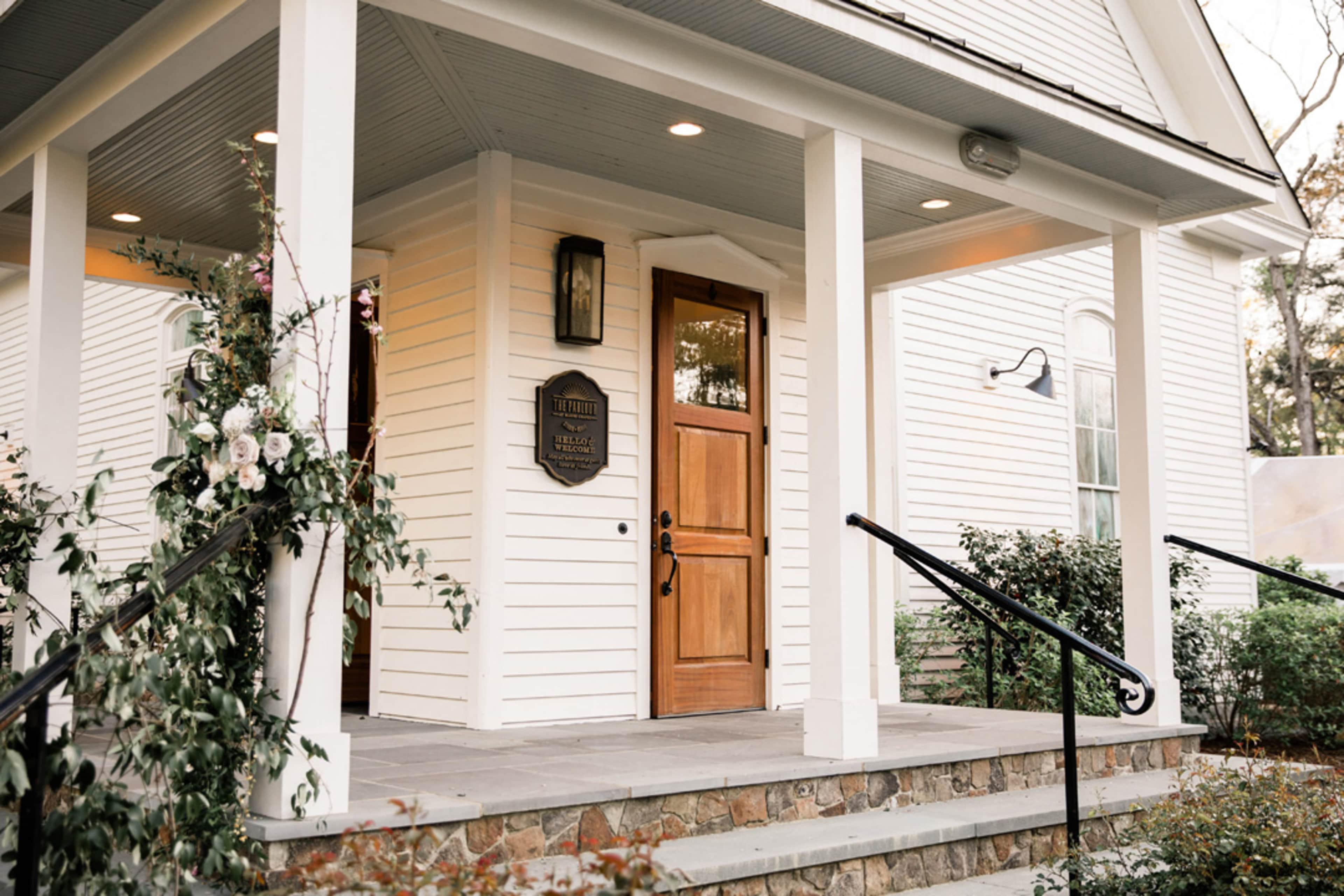 A white house with a wooden front door and a porch decorated with greenery and flowers.