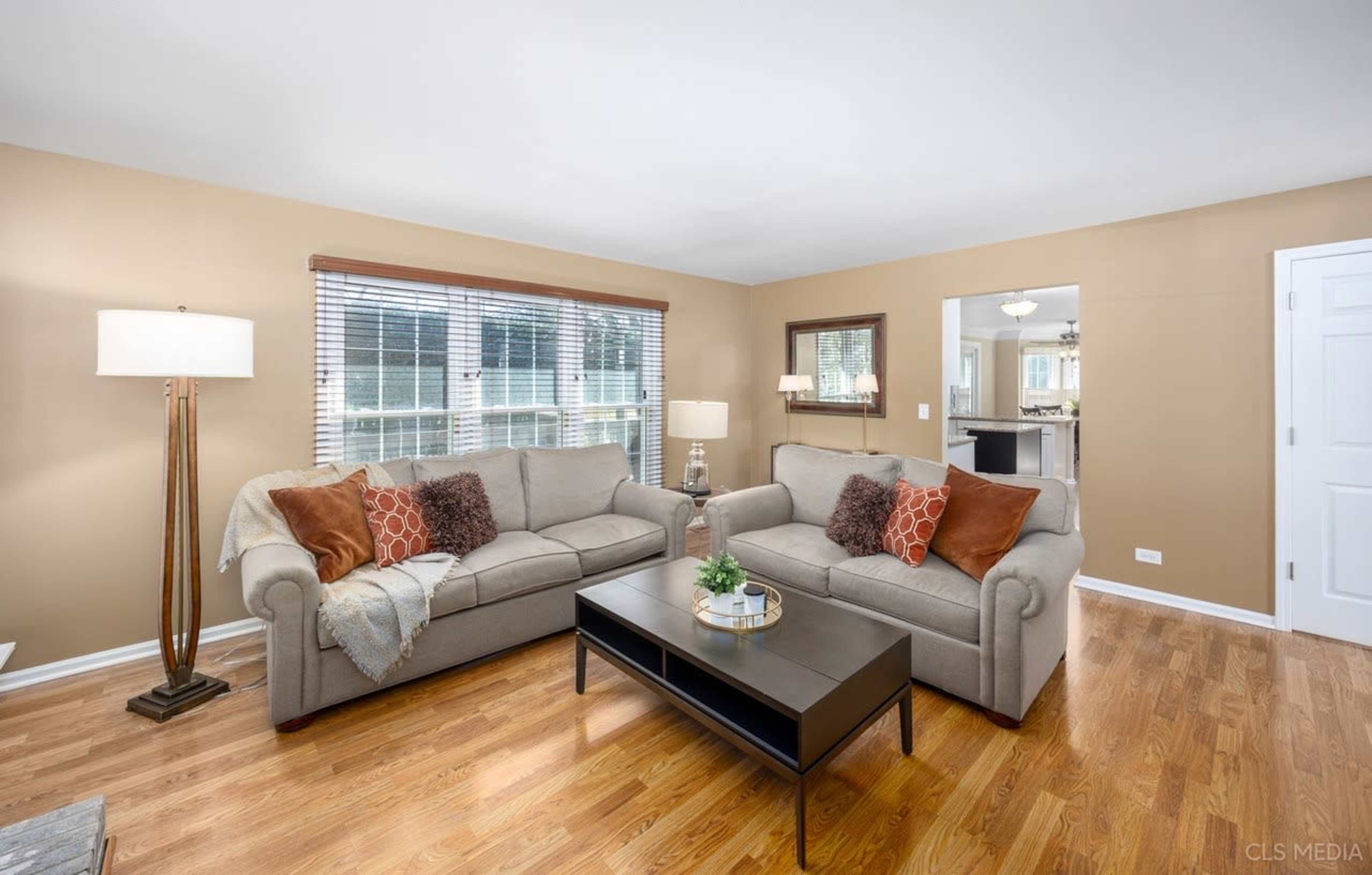 The living room features two sofas, a coffee table, and large windows with blinds, all set against a warm-toned wall.