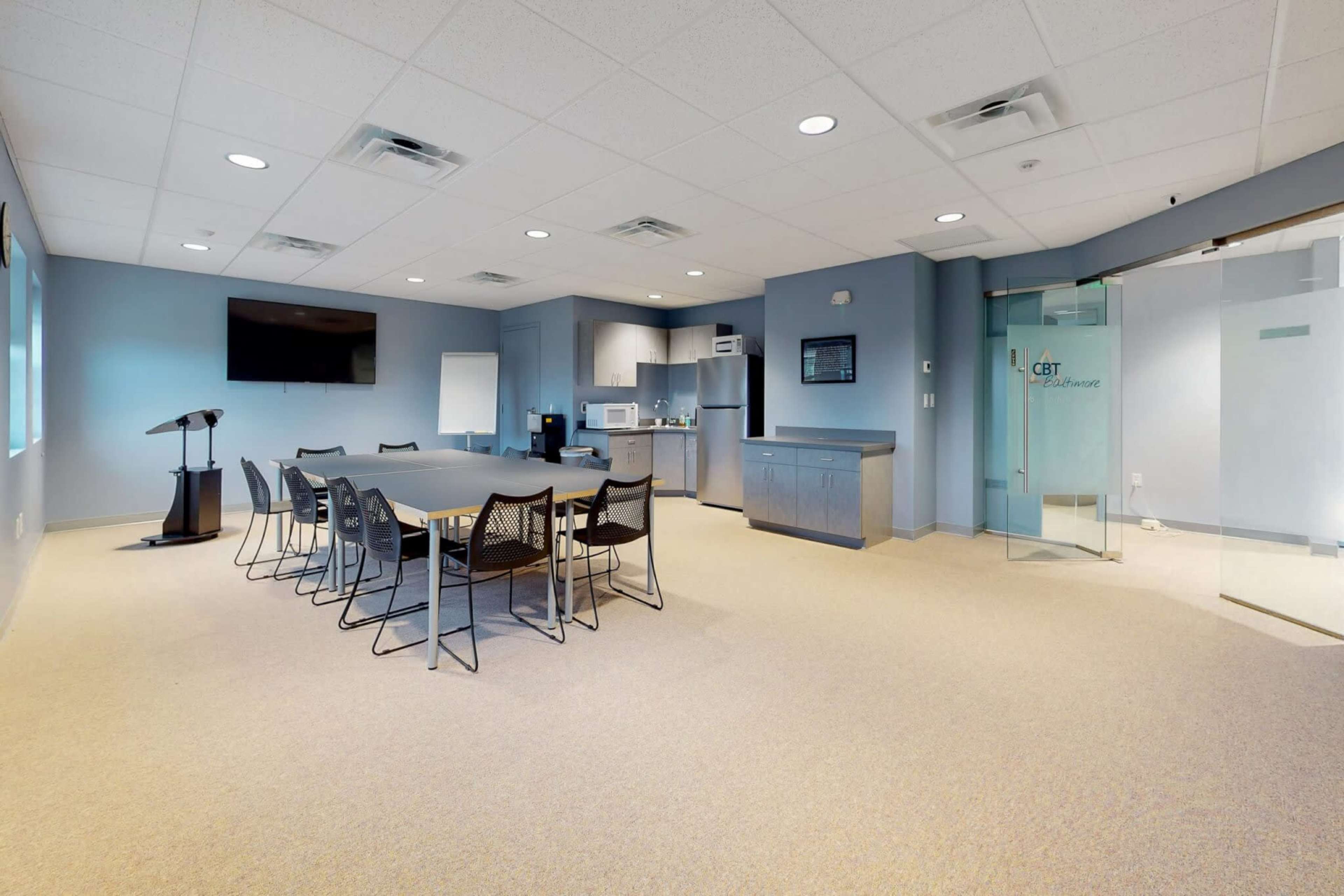 A large, modern conference room with a long table surrounded by chairs, a kitchenette area, and a glass door leading to another room.