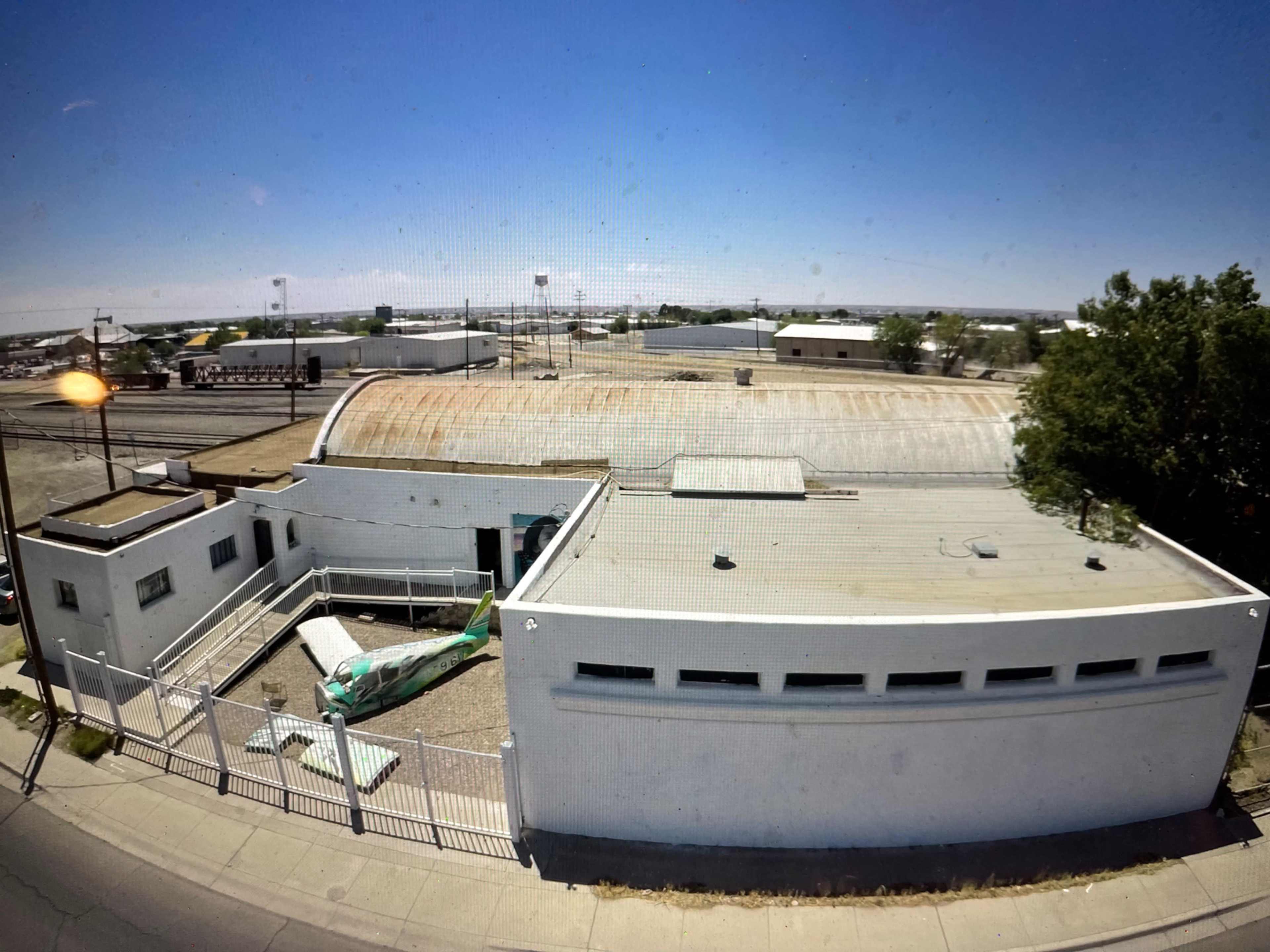 The image shows a flat-roofed white building with a green object in a gravel courtyard, surrounded by a metal fence, set against a clear blue sky and industrial structures in the background.