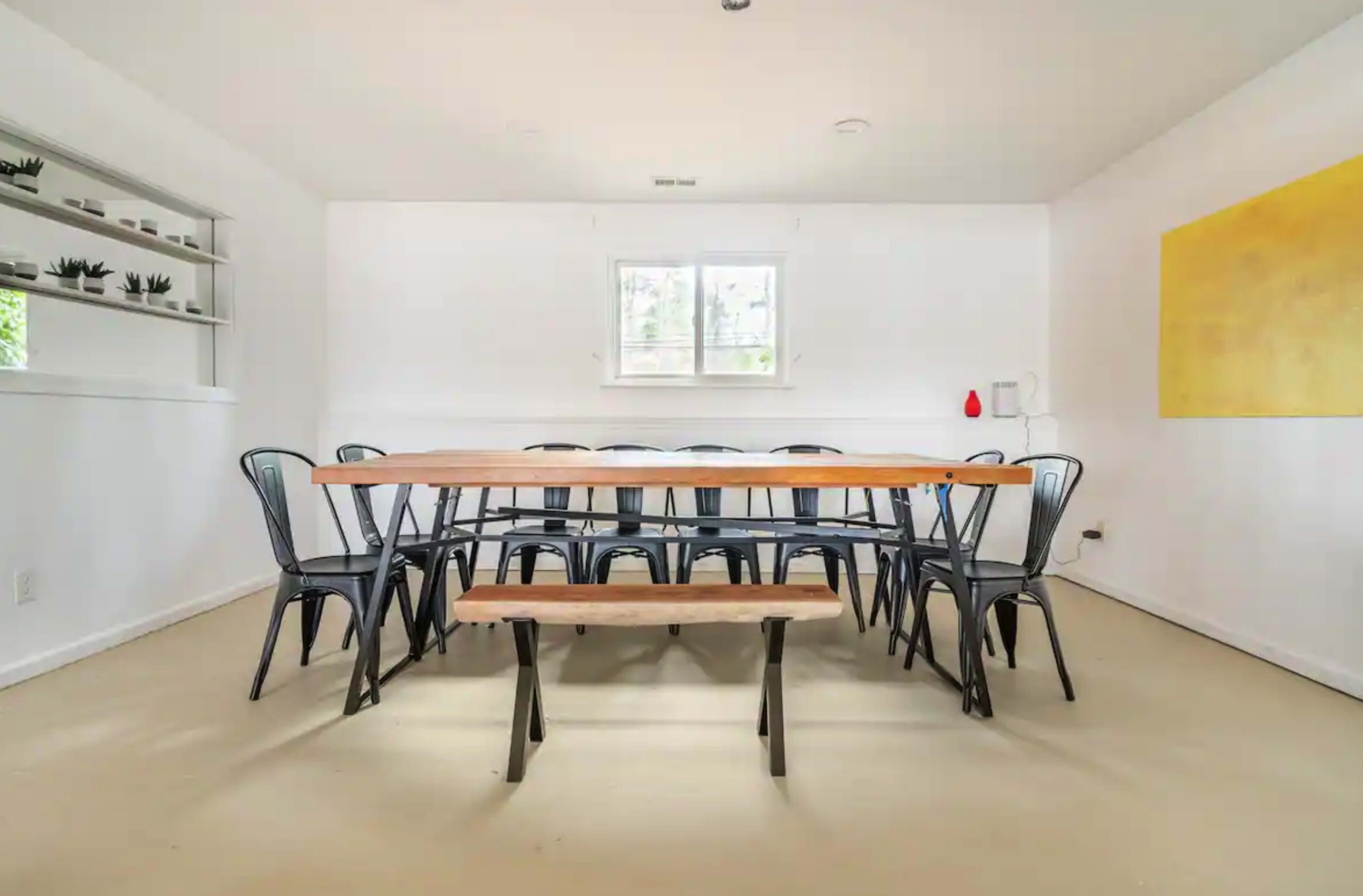 A spacious dining area featuring a long wooden table surrounded by black metal chairs, with a bench on one side and minimalist decor on the walls.