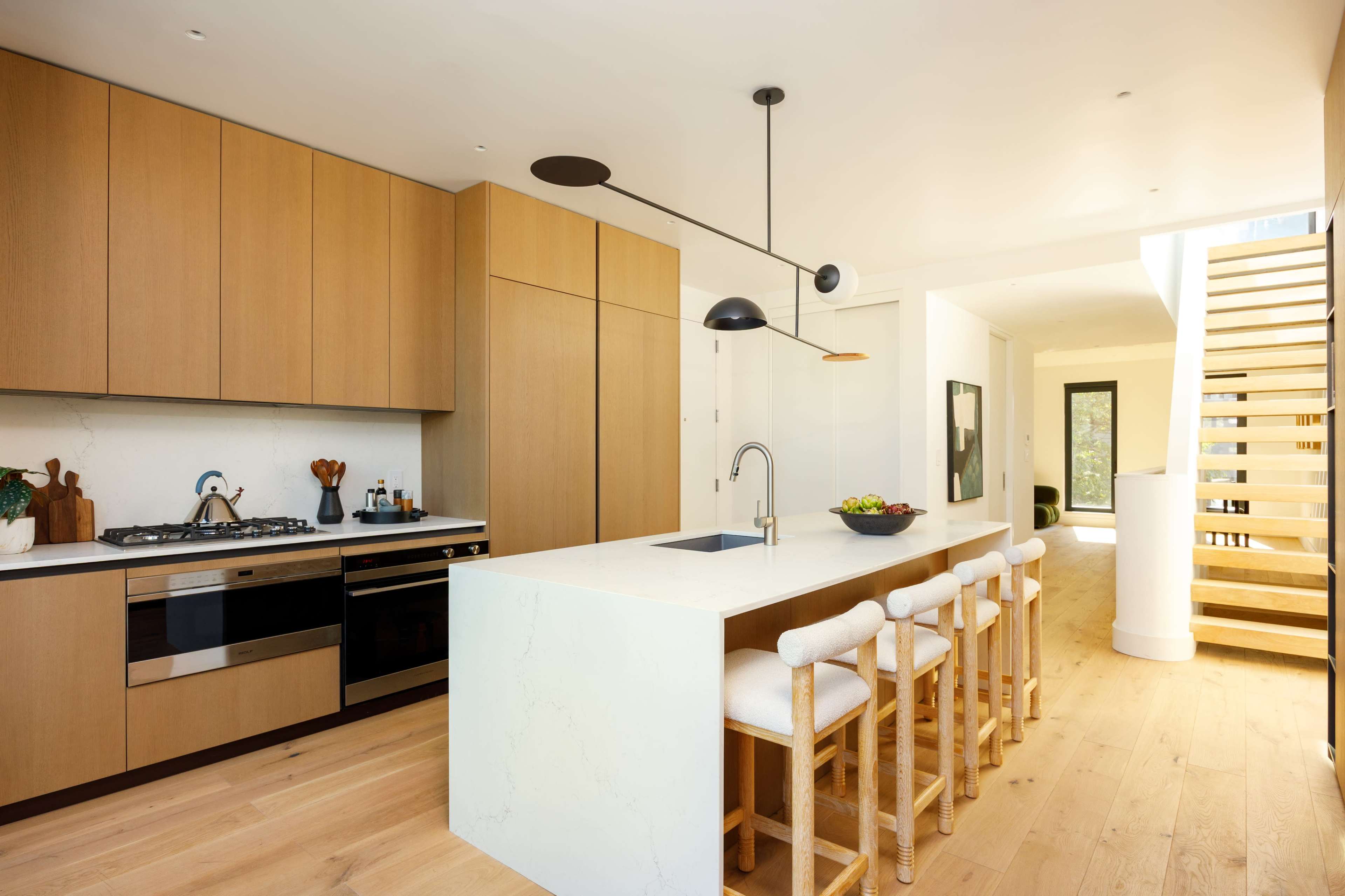 The image shows a modern kitchen with wooden cabinetry, a central island with seating, and a staircase leading to another area of the house.