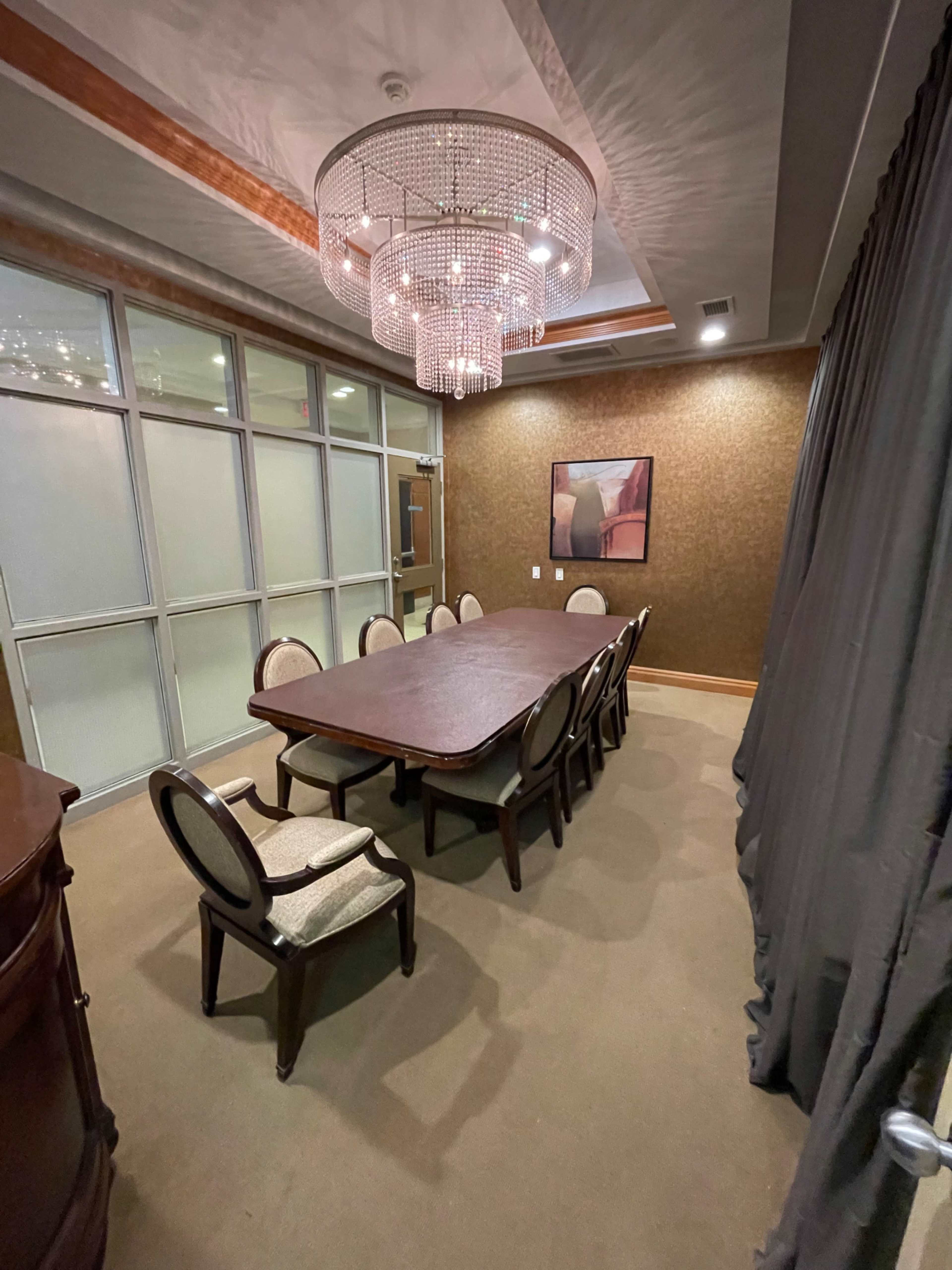 A conference room with a large wooden table surrounded by chairs, a chandelier overhead, and a wall covered in textured fabric.