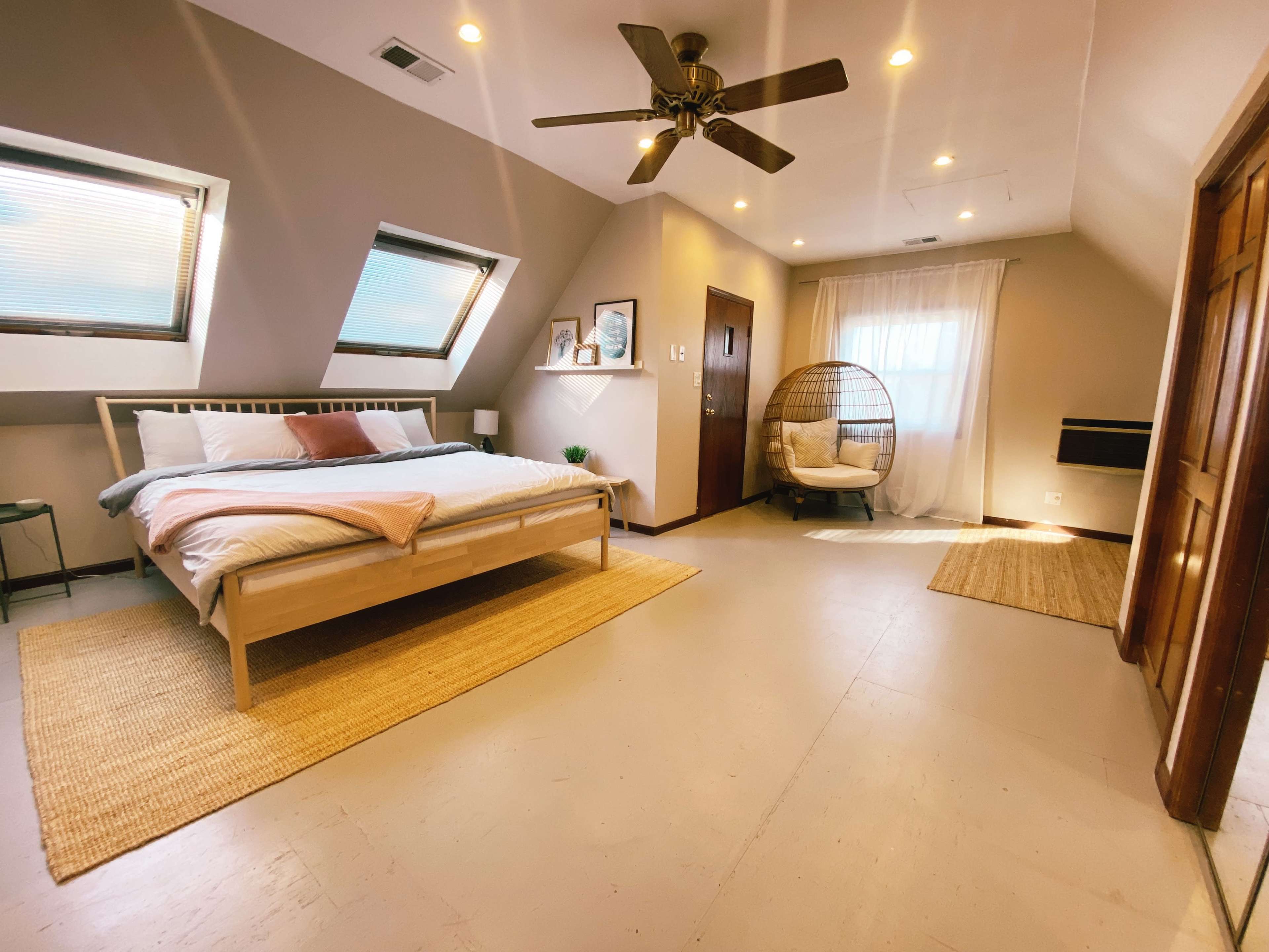 The image shows a well-lit attic bedroom featuring a bed, a rattan chair, and large windows with light curtains.