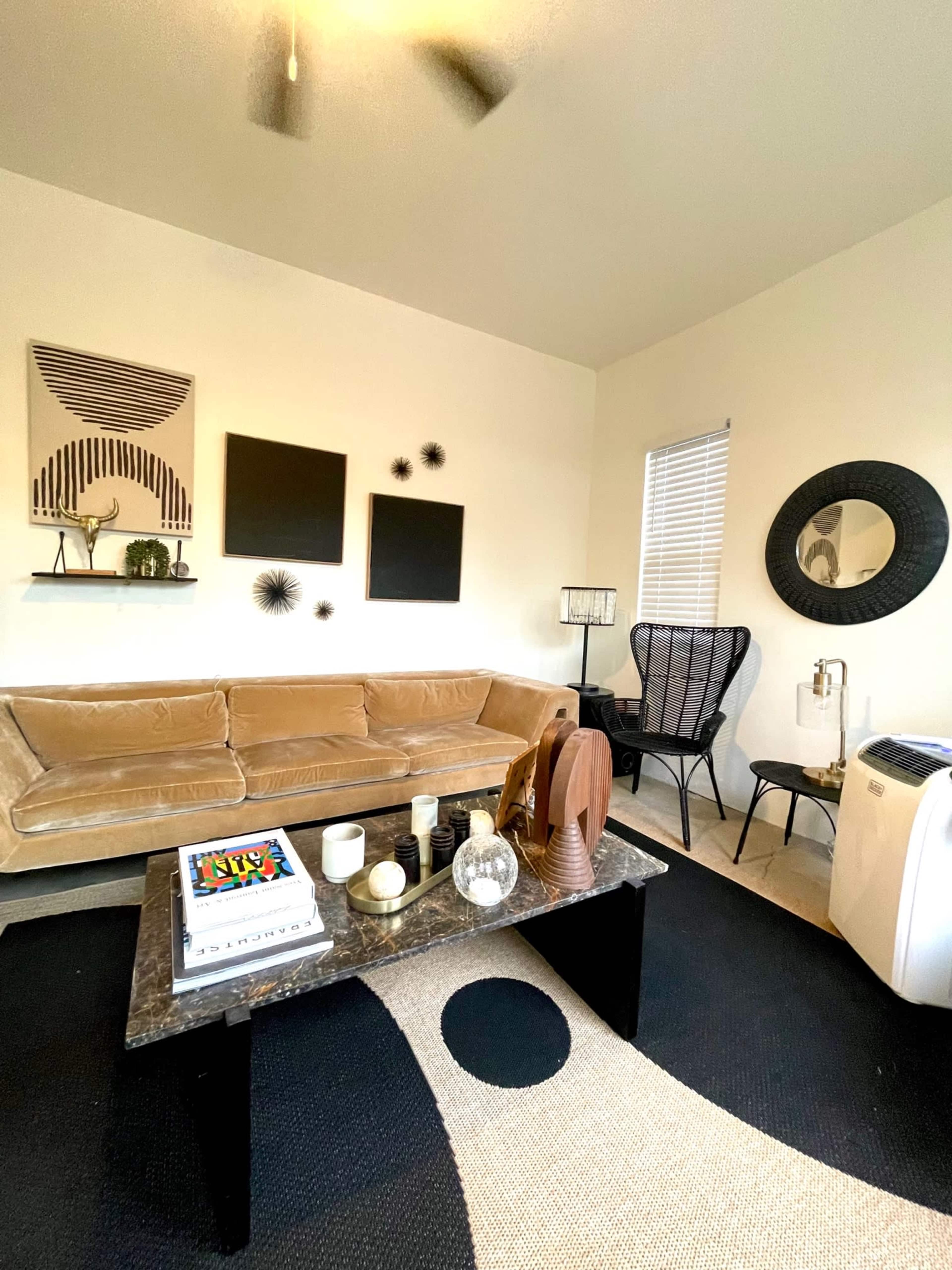 A living room with a beige sectional sofa, a black coffee table, decorative wall art, and a mix of furniture including a patterned chair and a round mirror.