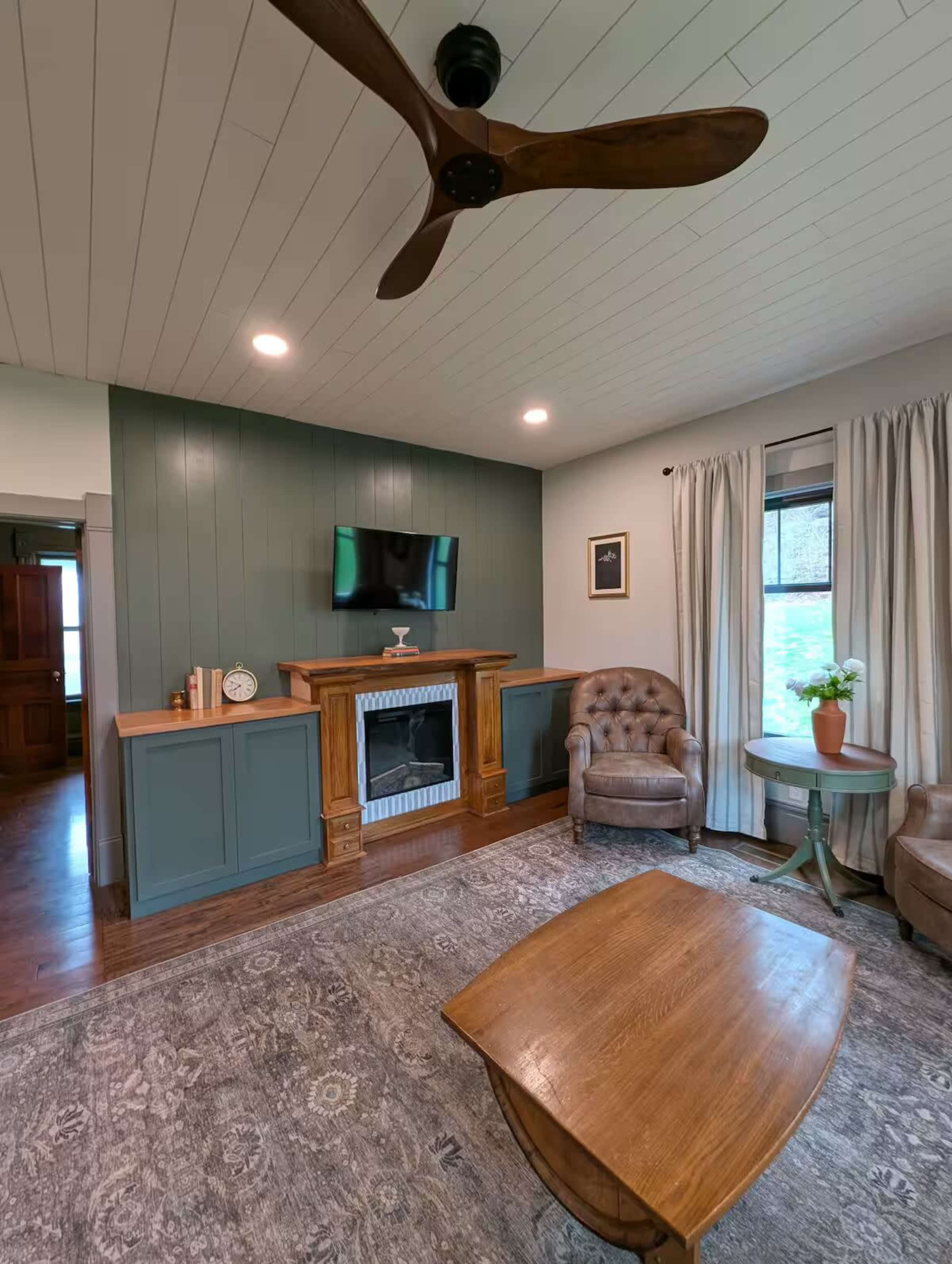 The image shows a living room with a fireplace, a flat-screen TV mounted on the wall, two armchairs, a wooden coffee table, and a window with natural light.